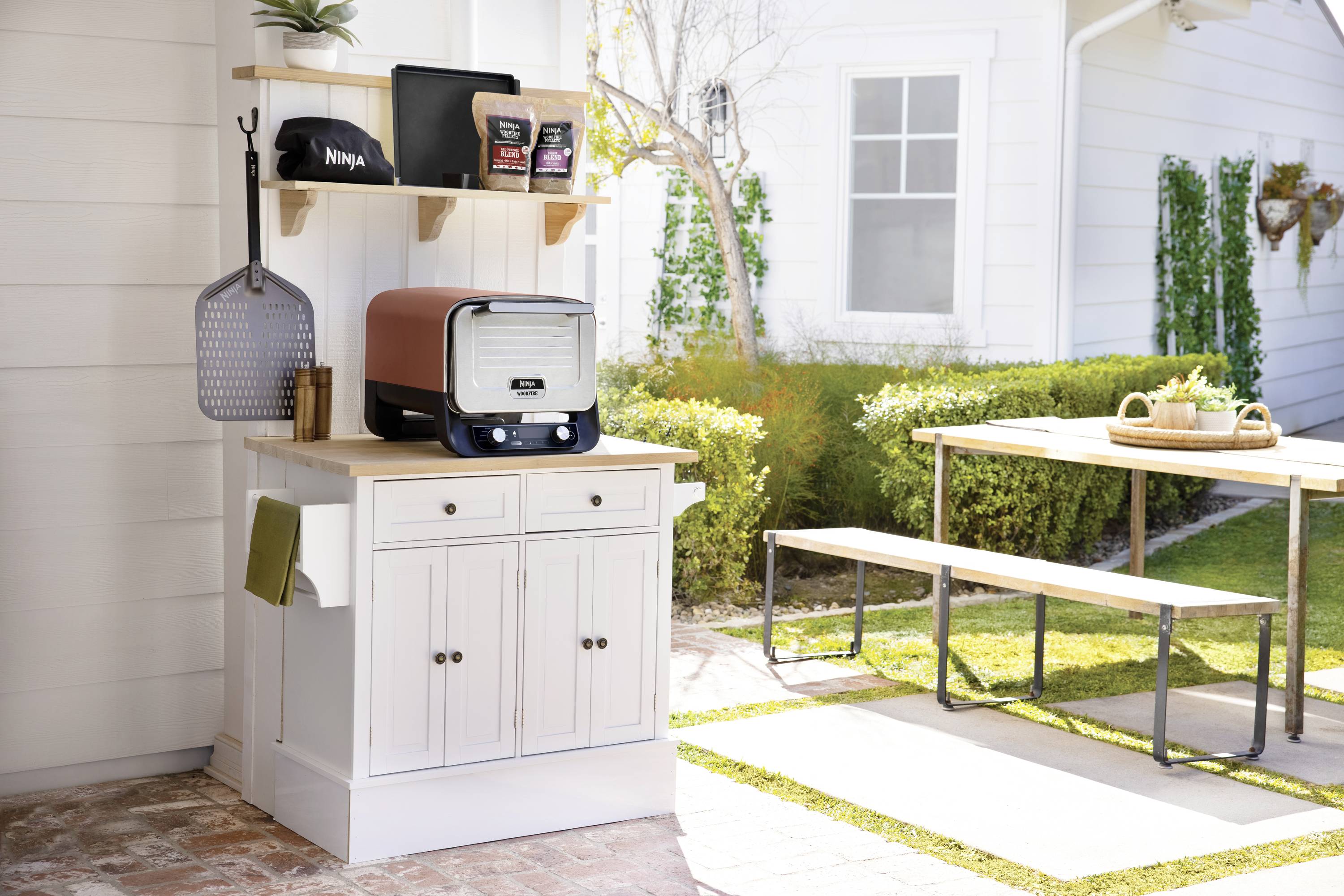 Egy pizzasütő egy fehér szekrényen a kertben. Mellette egy pizzalapát és egyéb eszközök lógnak. Egy asztal paddal zöld környezetben áll.