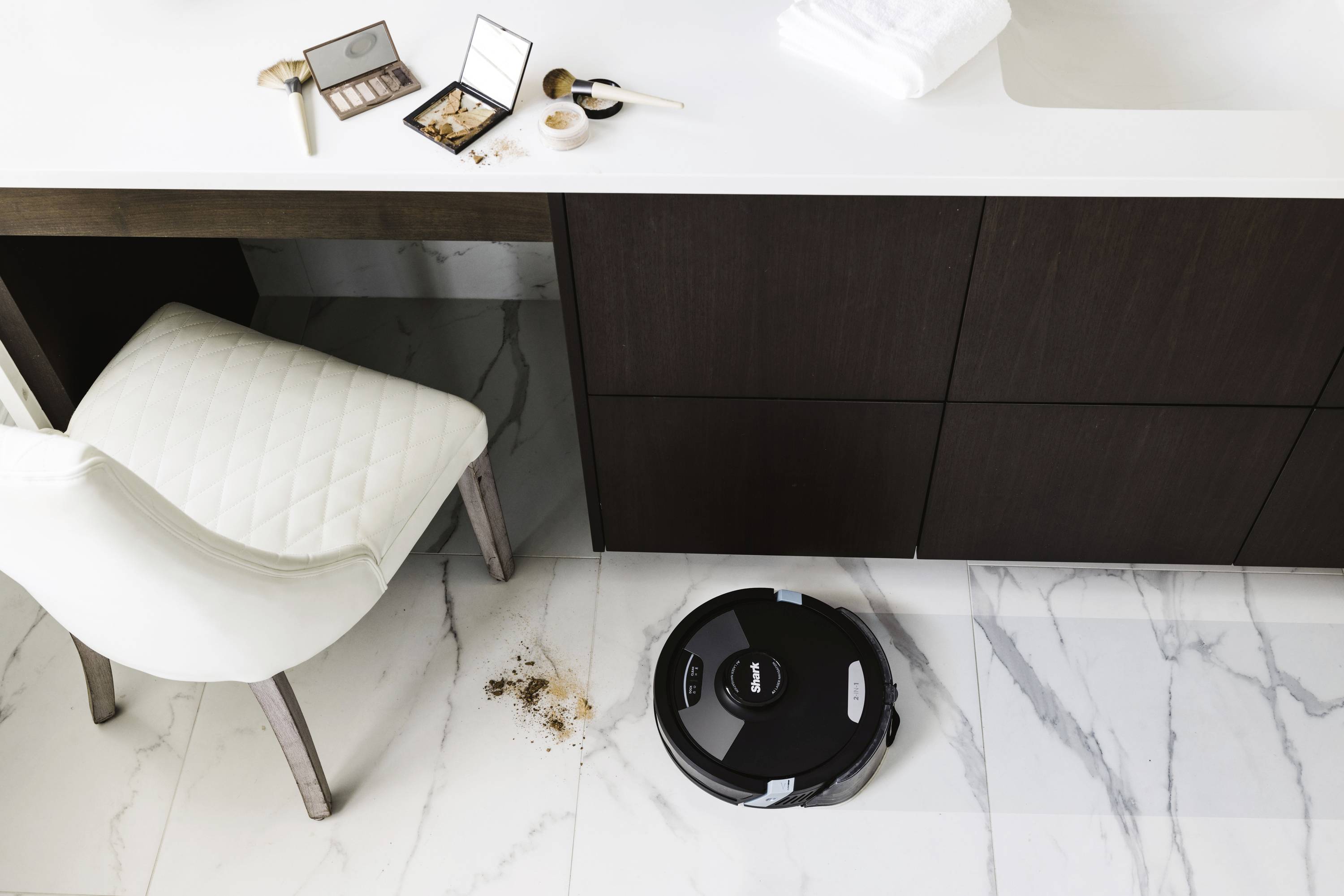 Egy robotporszívó takarítja a padlót egy modern mosdópult alatt. Kozmetikai termékek vannak szétszórva az asztalon. Tiszta, minimalista fürdőszoba.