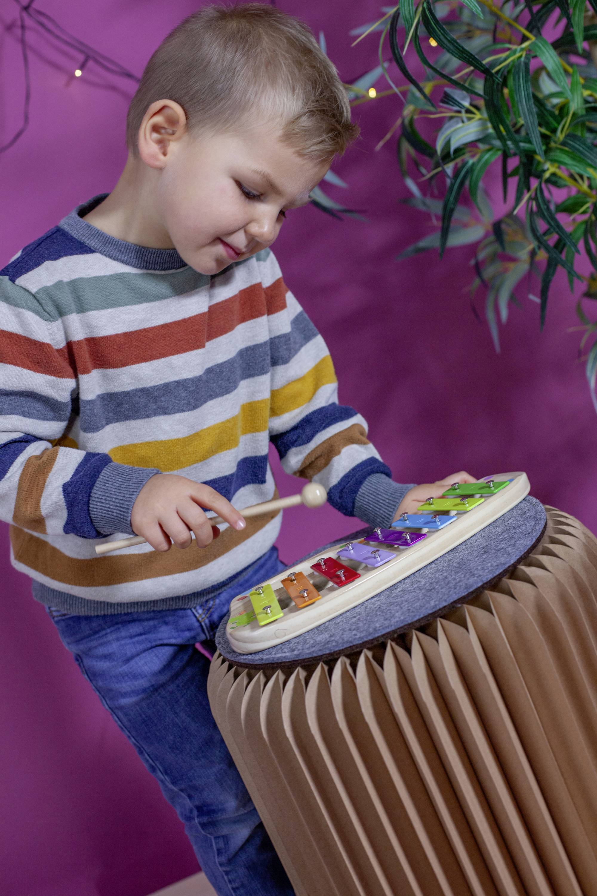 Egy gyerek egy fa ütővel játszik egy színes xilofónon, amely egy kerek asztalon áll. A háttérben növények láthatók.