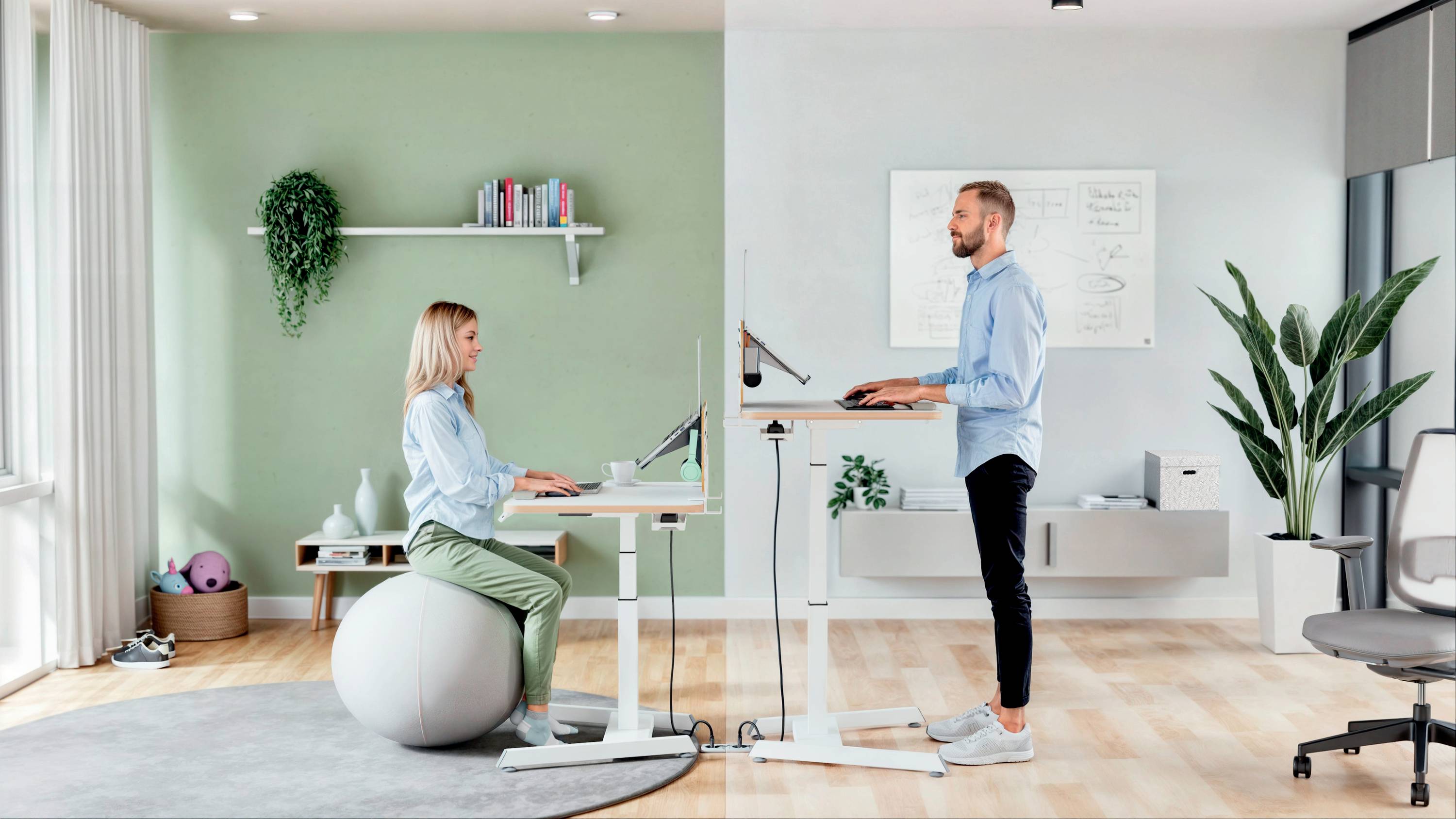 Egy nő és egy férfi egy ergonomikus irodában dolgozik. Ő egy gimnasztikai labdán ül, ő pedig egy magasságállítható íróasztal mellett áll.