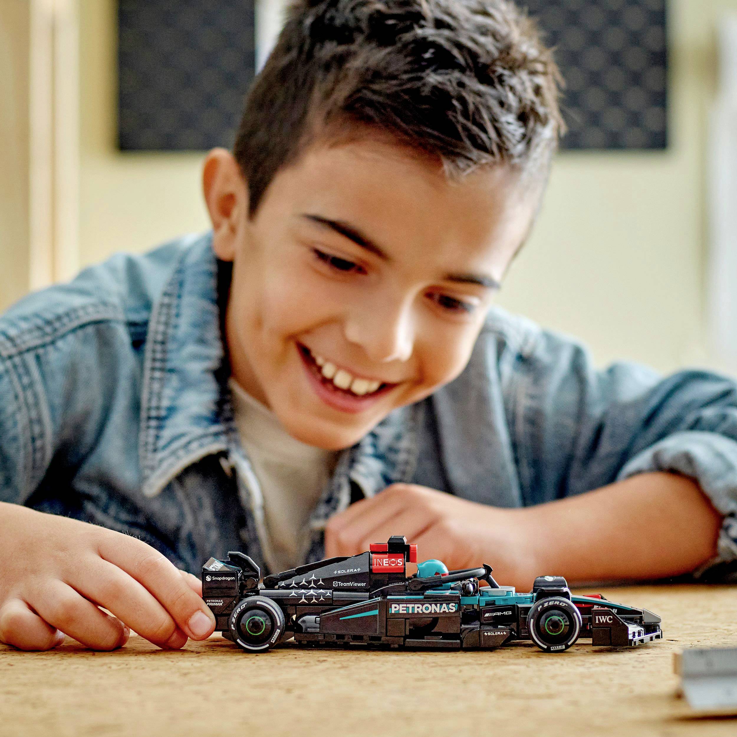 Egy gyerek mosolyog és egy versenyautó stílusú modellautóval játszik egy asztalnál.
