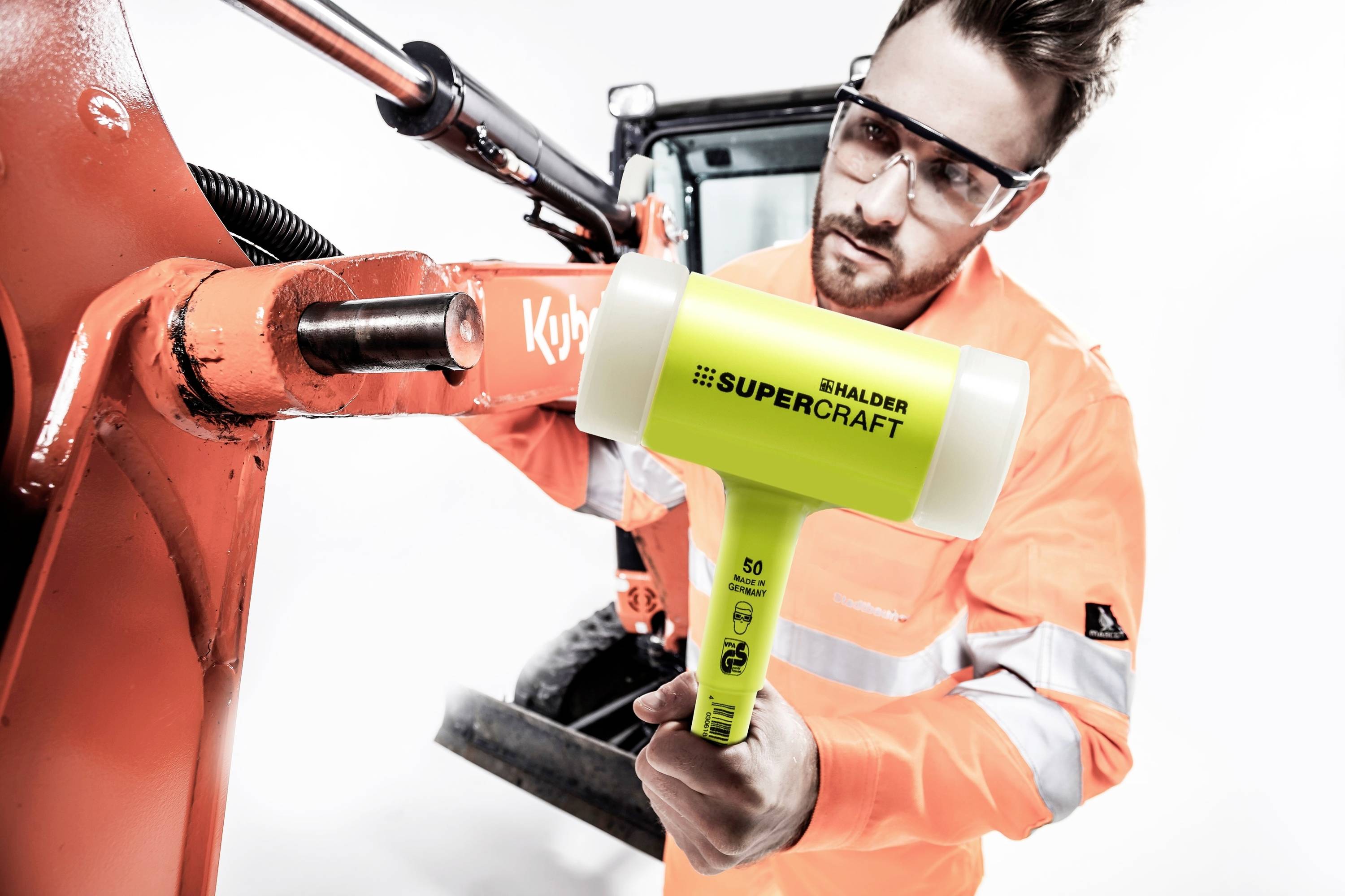 Egy férfi védőruházatban egy sárga Halder védőkalapácsot használ egy narancssárga gépalkatrészen végzett munkájához.
