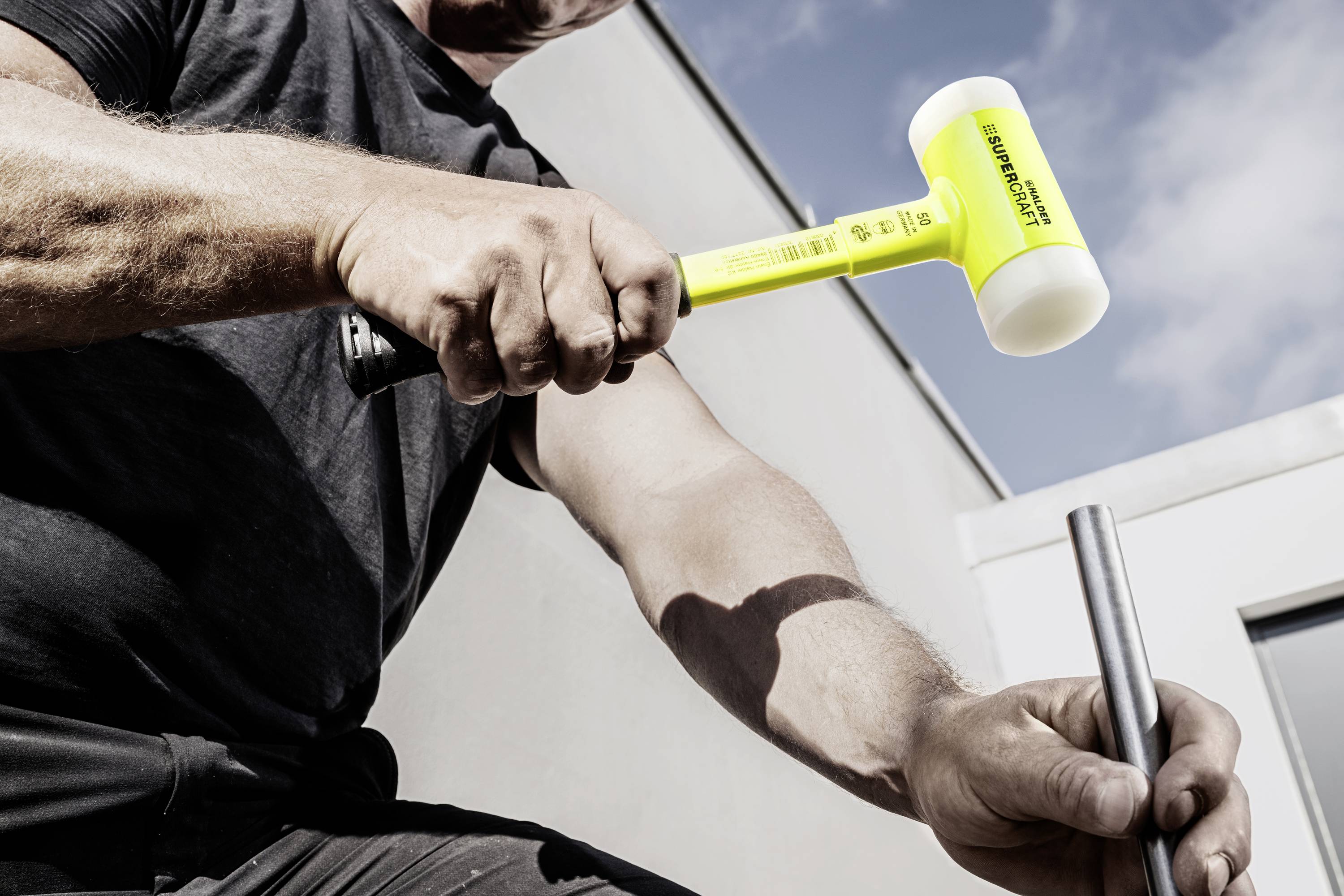 Egy férfi egy sárga védőkalapáccsal üt egy fémrudat. Az ég tiszta és kék a háttérben.