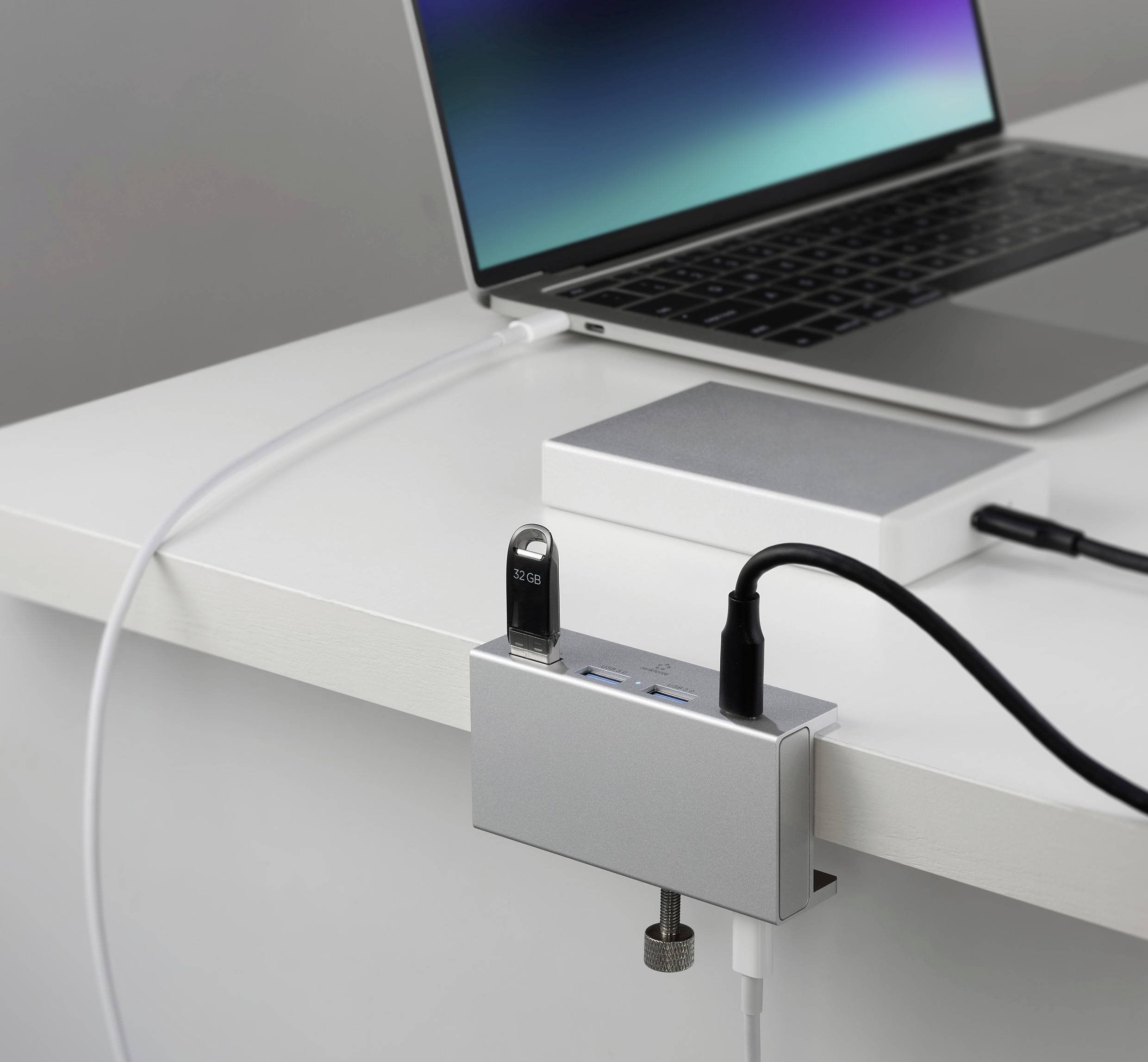 Egy laptop nyitott képernyővel áll egy asztalon. Egy USB-elosztó csatlakoztatva van, és egy USB-pendrive és kábelek vannak bedugva.