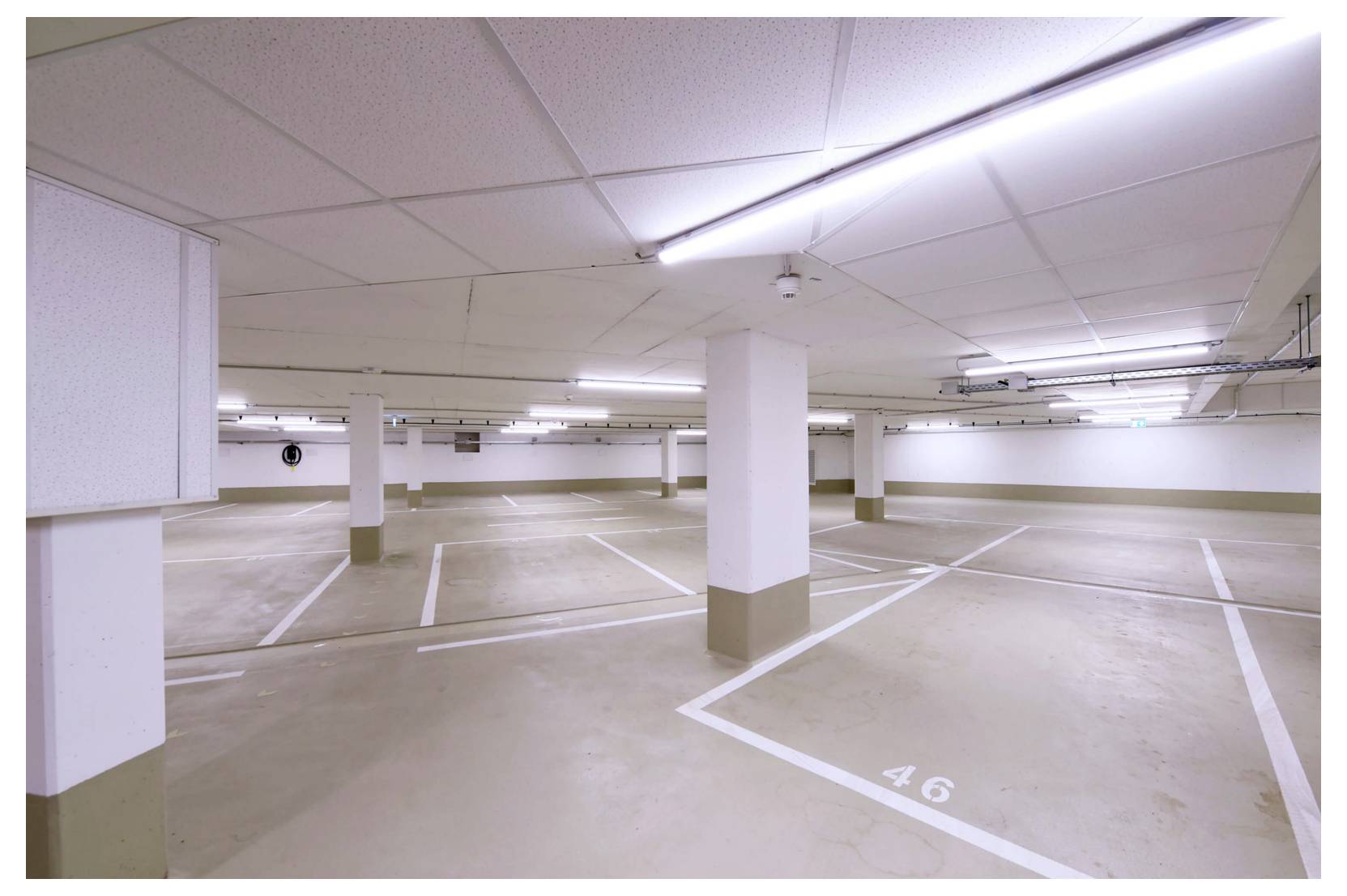Üres beltéri parkoló garázs számozott parkolóhelyekkel és fehér tartoszlopokkal, fénycsöves mennyezeti lámpák alatt.