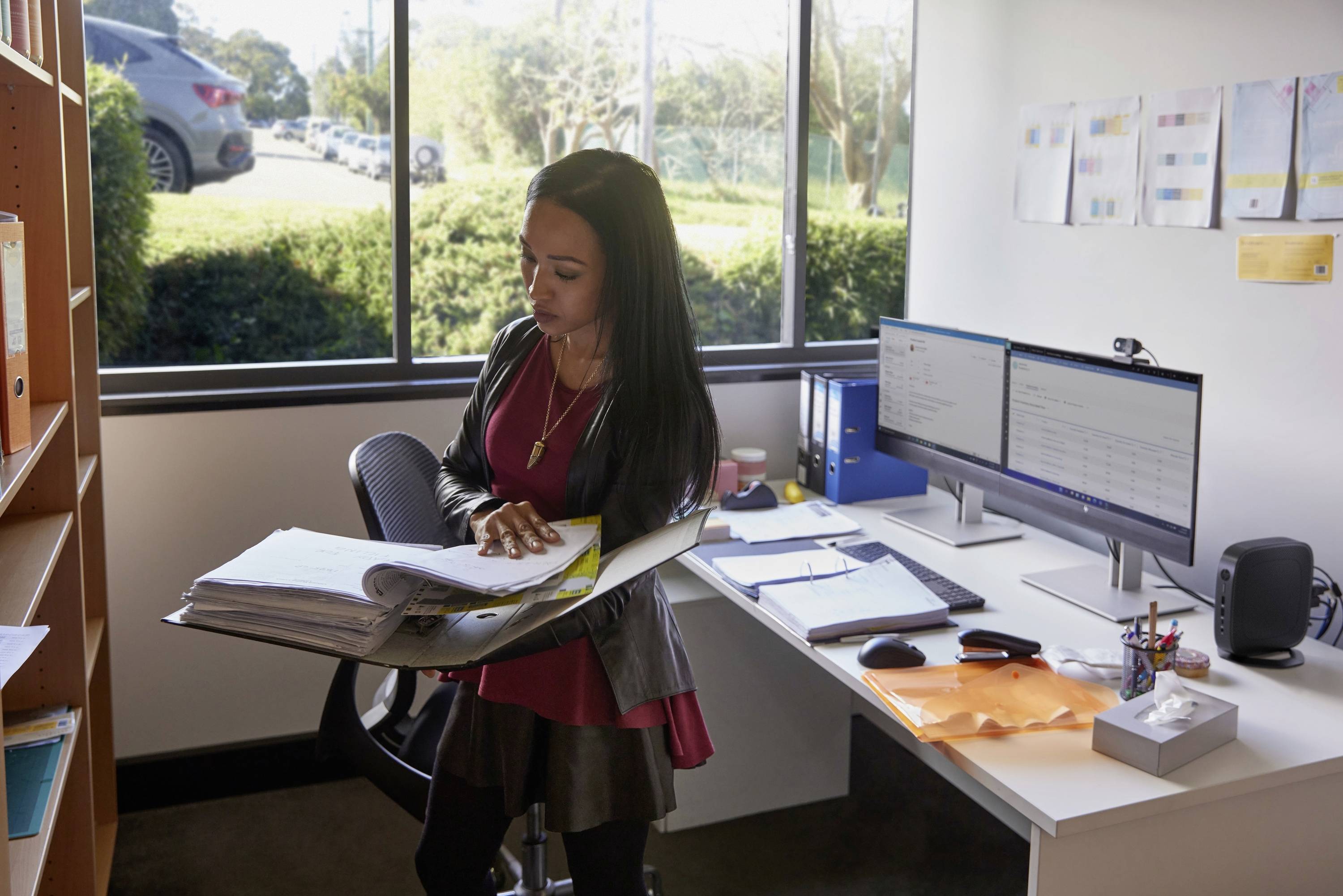 Egy személy áll egy irodában egy állópultnál, és iratokat ellenőriz. A háttérben egy íróasztal két monitorral és egy ablak látható.