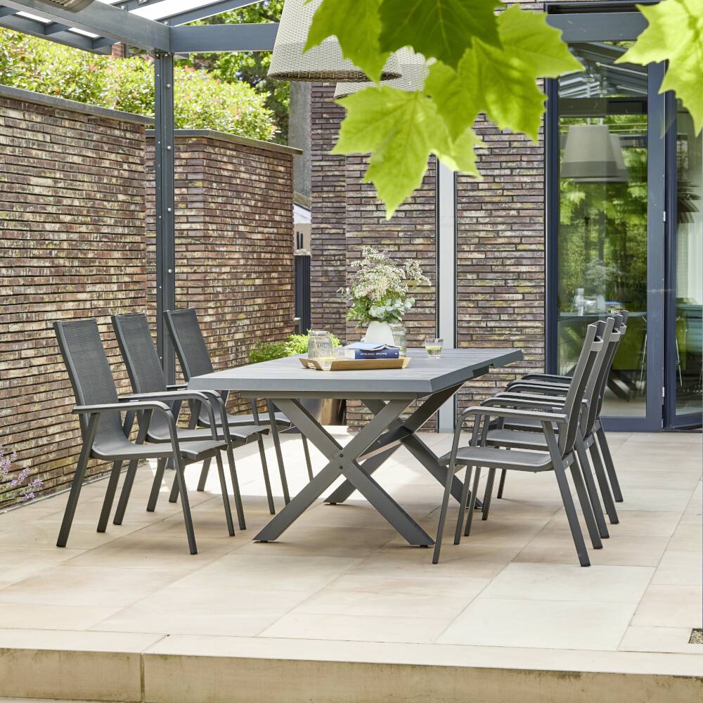 Terasz modern szürke fémszékekkel és asztallal egy téglfalakkal és üvegtetővel körülvett kertben, zöld növényzet által övezve.