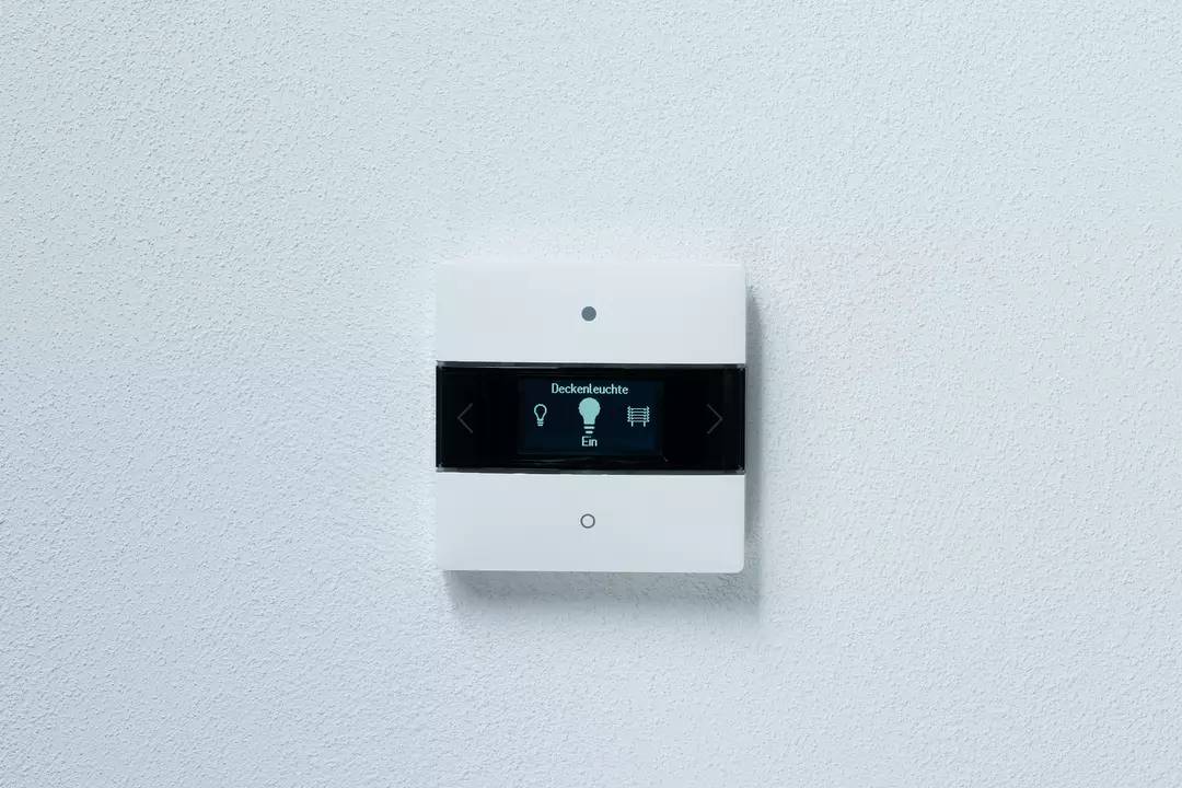 Egy fehér fali kapcsoló digitális kijelzővel mutatja a 'Mennyezeti lámpa bekapcsolva' feliratot és a világításvezérlés szimbólumait.