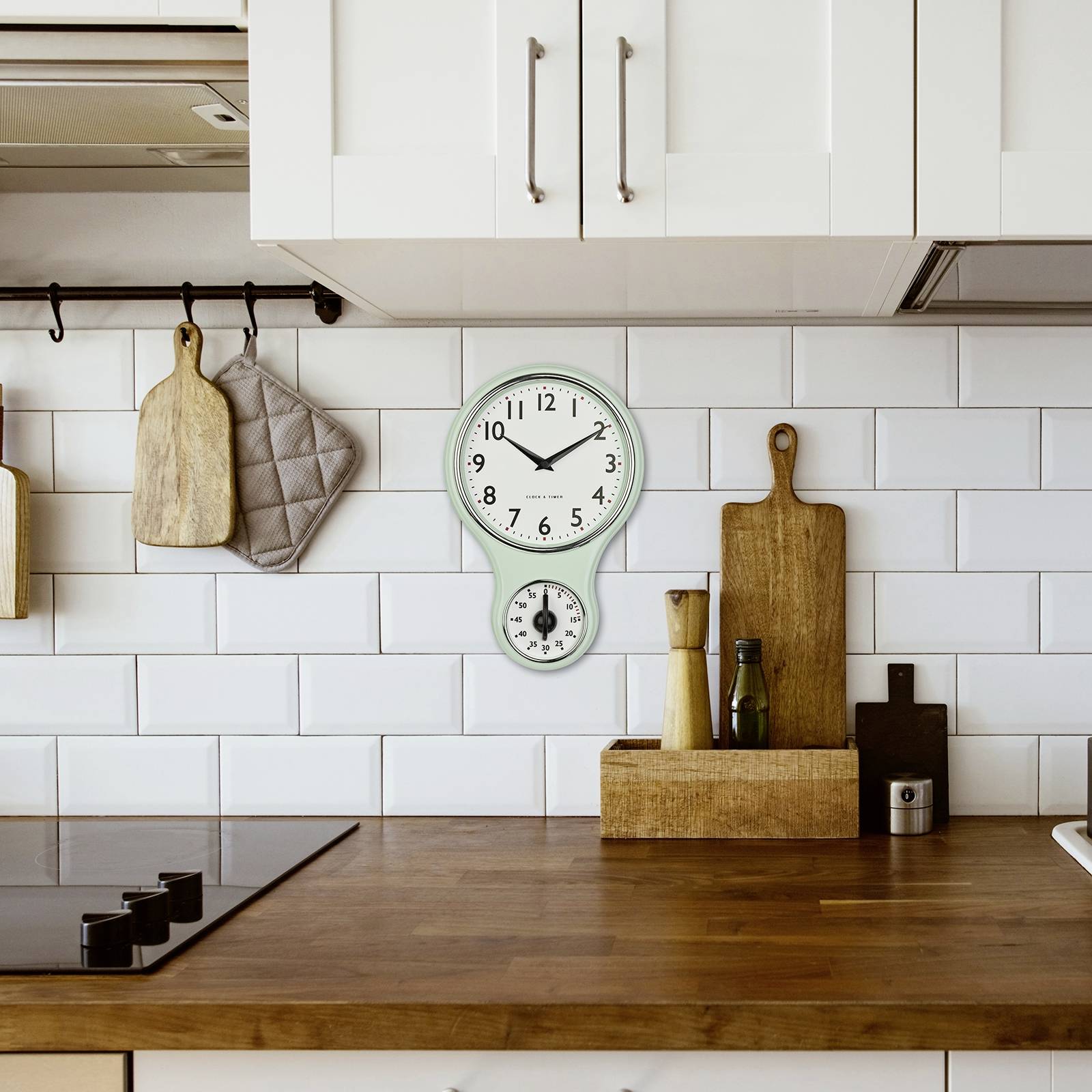 Egy konyha fehér csempézett falakkal, fa munkalappal, vágódeszkákkal, fűszerekkel és egy fali órával, amely 10:09-et mutat.