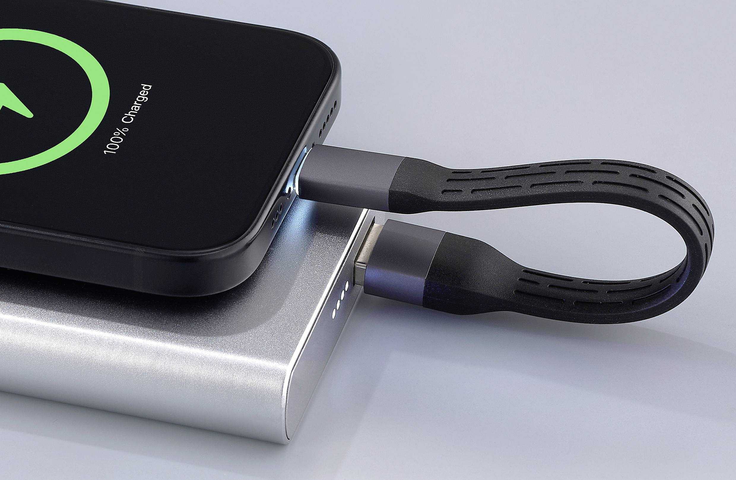 Egy okostelefon egy fekete kábel segítségével tölt egy ezüst színű powerbank-ről. A képernyőn '100% feltöltve' felirat látható.