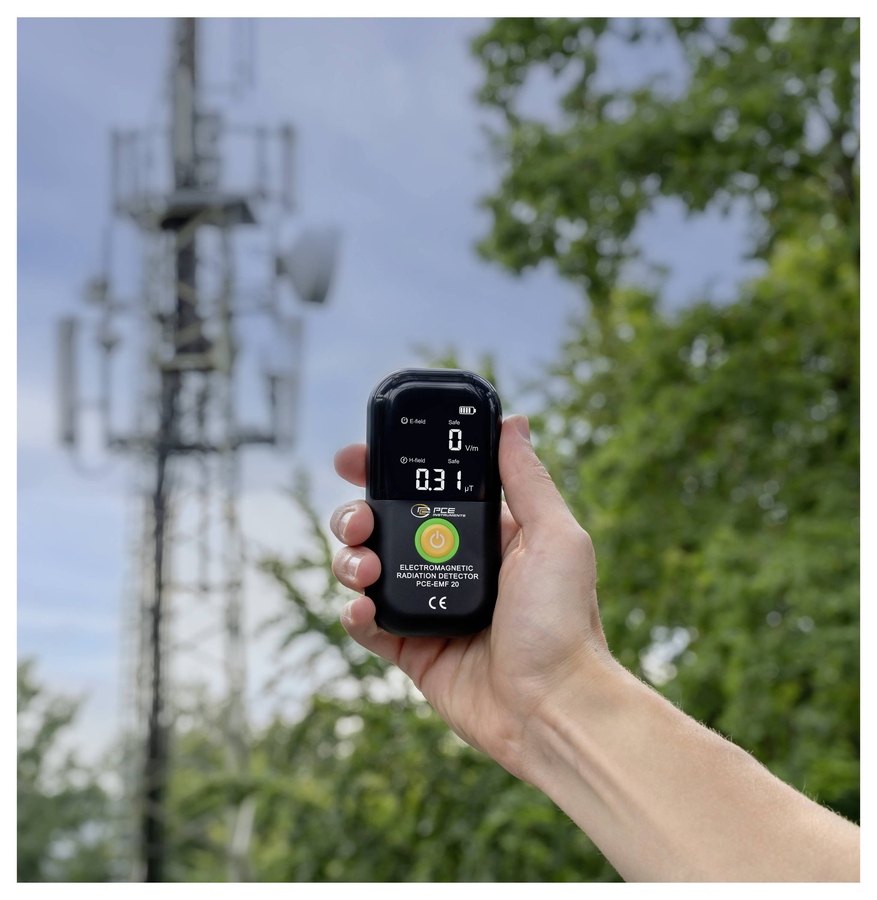 Egy kéz digitális elektromágneses sugárzásdetektort tart a szabadban egy mobiltelefon-torony közelében, a képernyőn egy kis numerikus leolvasás látható.