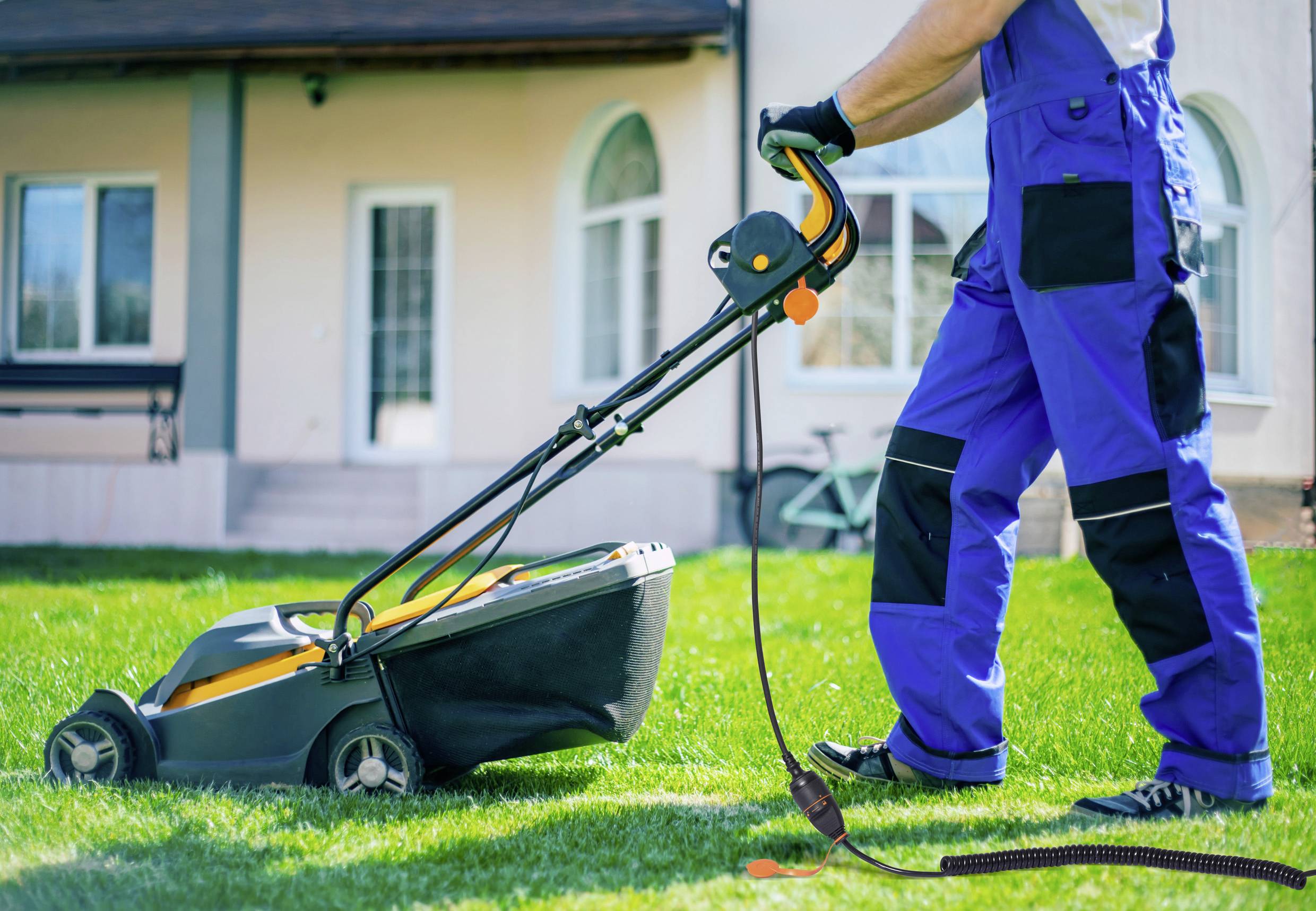 Egy személy elektromos fűnyíróval nyírja a füvet; kék munkaruhát visel. A háttérben egy ház látható.
