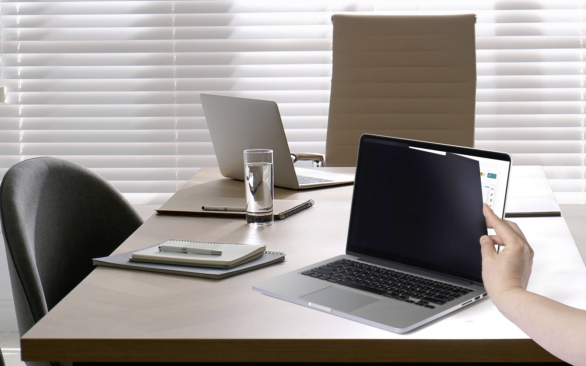 Egy iroda három laptoppal az asztalon, mellettük egy pohár víz és íróeszközök. Egy személy egy laptopképernyőt tart.