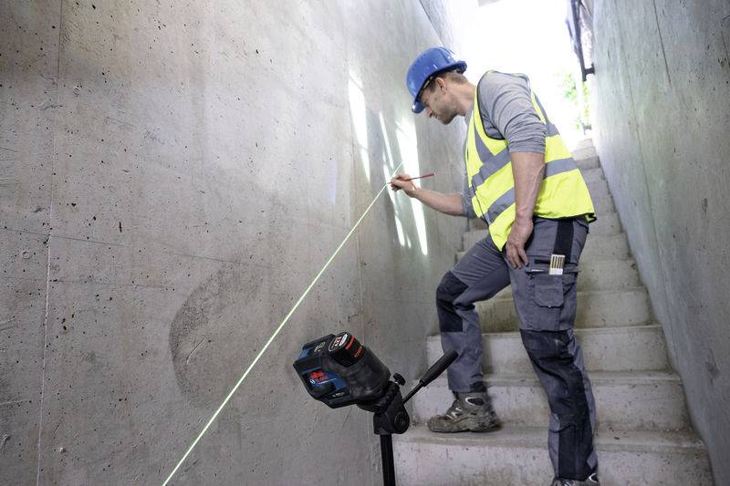 Egy védősisakot és fényvisszaverő mellényt viselő személy egy lézer-szintező készülékkel von egy vonalat egy betonfalon.