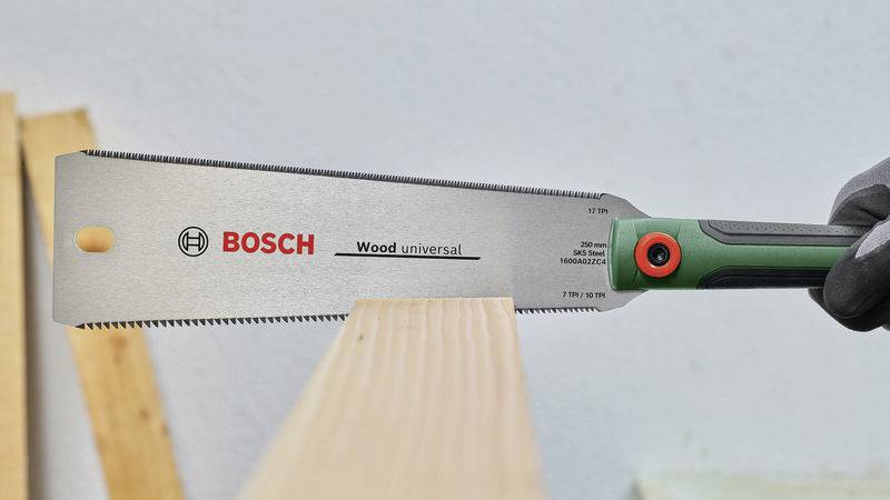 Egy személy egy Bosch Wood Universal fűrésszel vág egy fadeszkát. A fűrész zöld markolattal rendelkezik és látható rajta a Bosch logó.