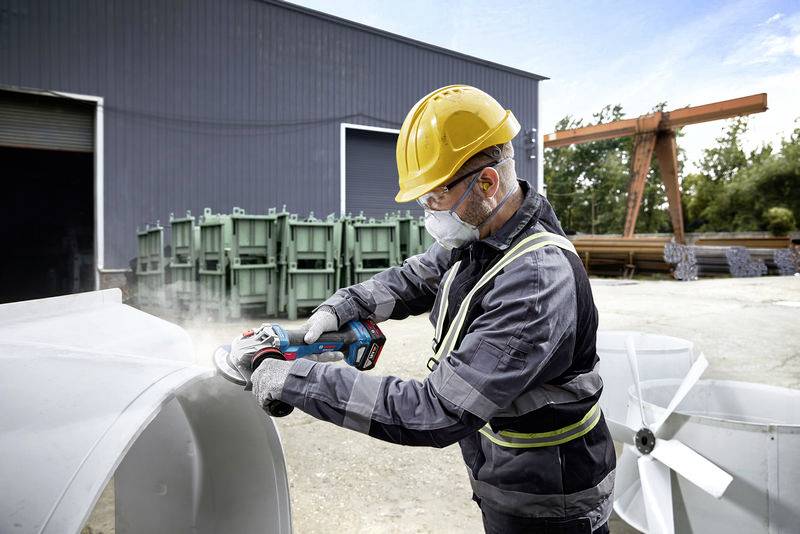 Egy védősisakot és maszkot viselő személy egy nagy fémdarabot csiszol egy ipari területen. A háttérben raktárépületek láthatók.