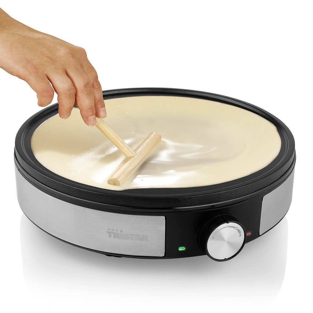 Egy kéz folyékony tésztát oszlat el egy kerek, fűtött palacsintasütőn egy falapáttal.