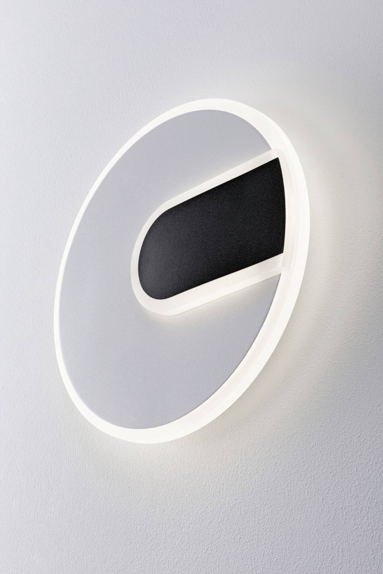 Kerek, fehér fali lámpa fekete, ovális betéttel; minimalista design, fehér falra szerelve.