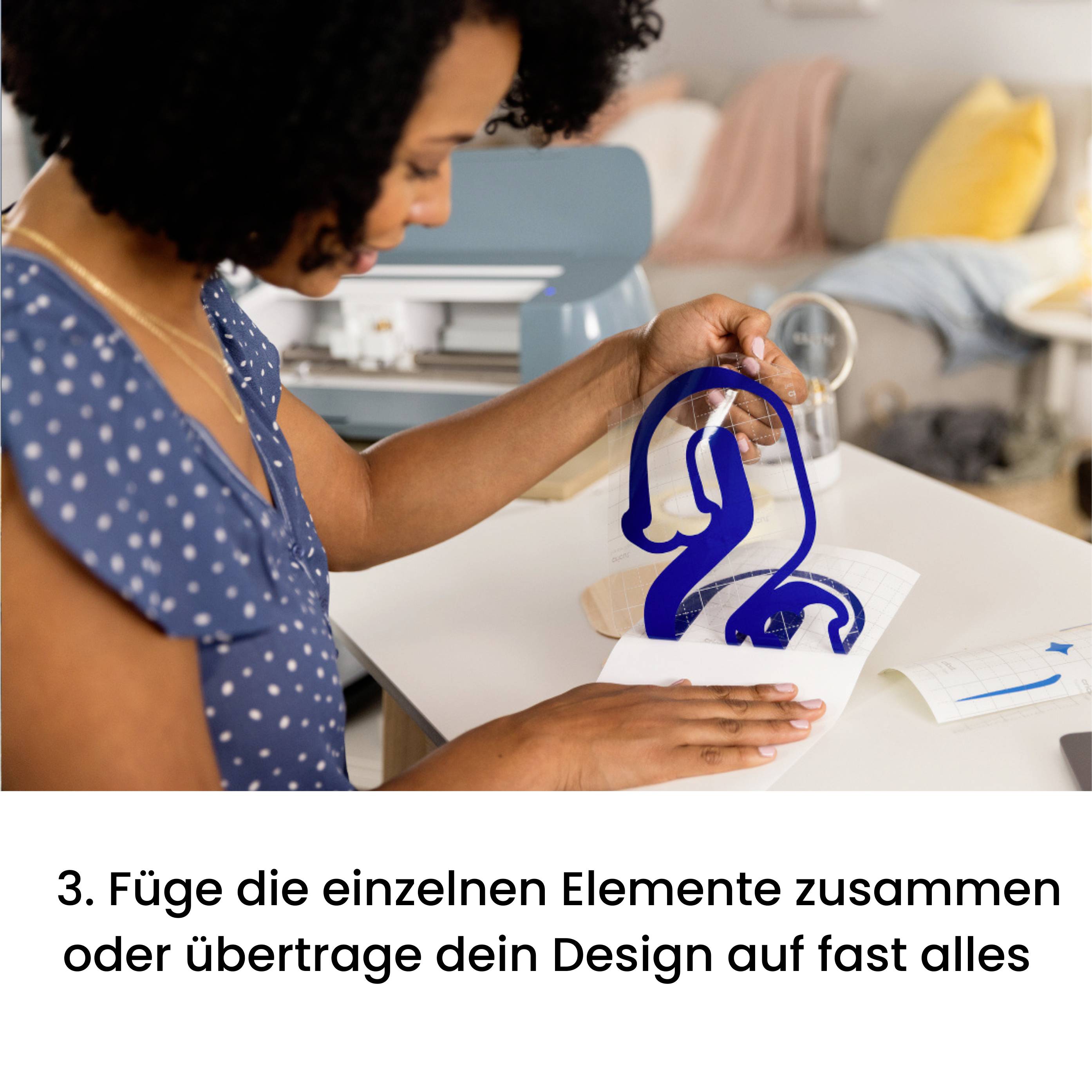 Egy személy kék designelemeket helyez fel egy vasalható képre egy asztalon. A háttérben egy nappali látható.