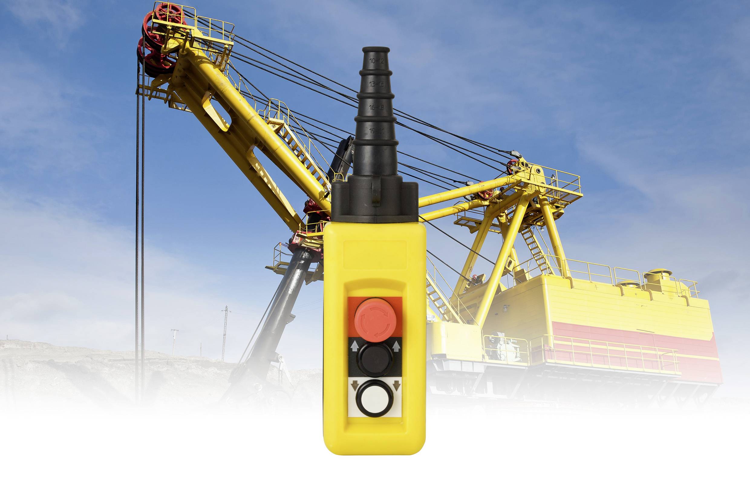 Kék ég alatt egy nagy, sárga kotrógép előtt egy sárga ipari távirányító gombok láthatók, amely a berendezés irányítását jelzi.