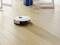 Egy fehér robotporszívó takarít egy fa padlón egy napfényes helyiségben. A háttérben asztalláb látható.