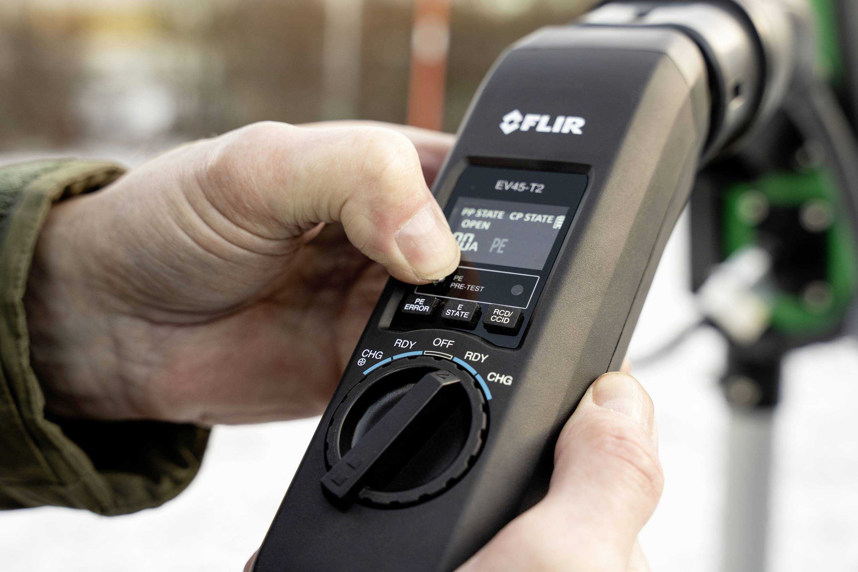 Egy személy kezel egy FLIR-mérőkészüléket, amely adatokat jelenít meg egy kijelzőn. A háttérben egy elmosódott terület látható.