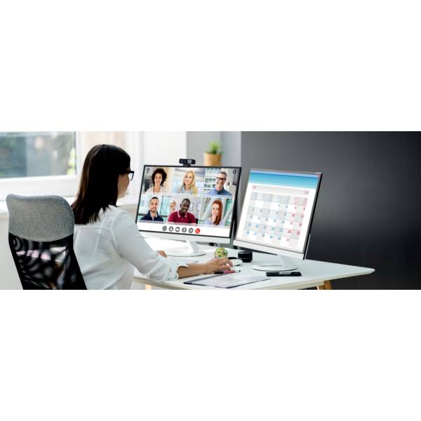 Egy nő ül az íróasztalánál és videokonferencián vesz részt két monitor előtt. Az egyik monitoron konferenciarésztvevők láthatók, a másikon naptárbejegyzések.