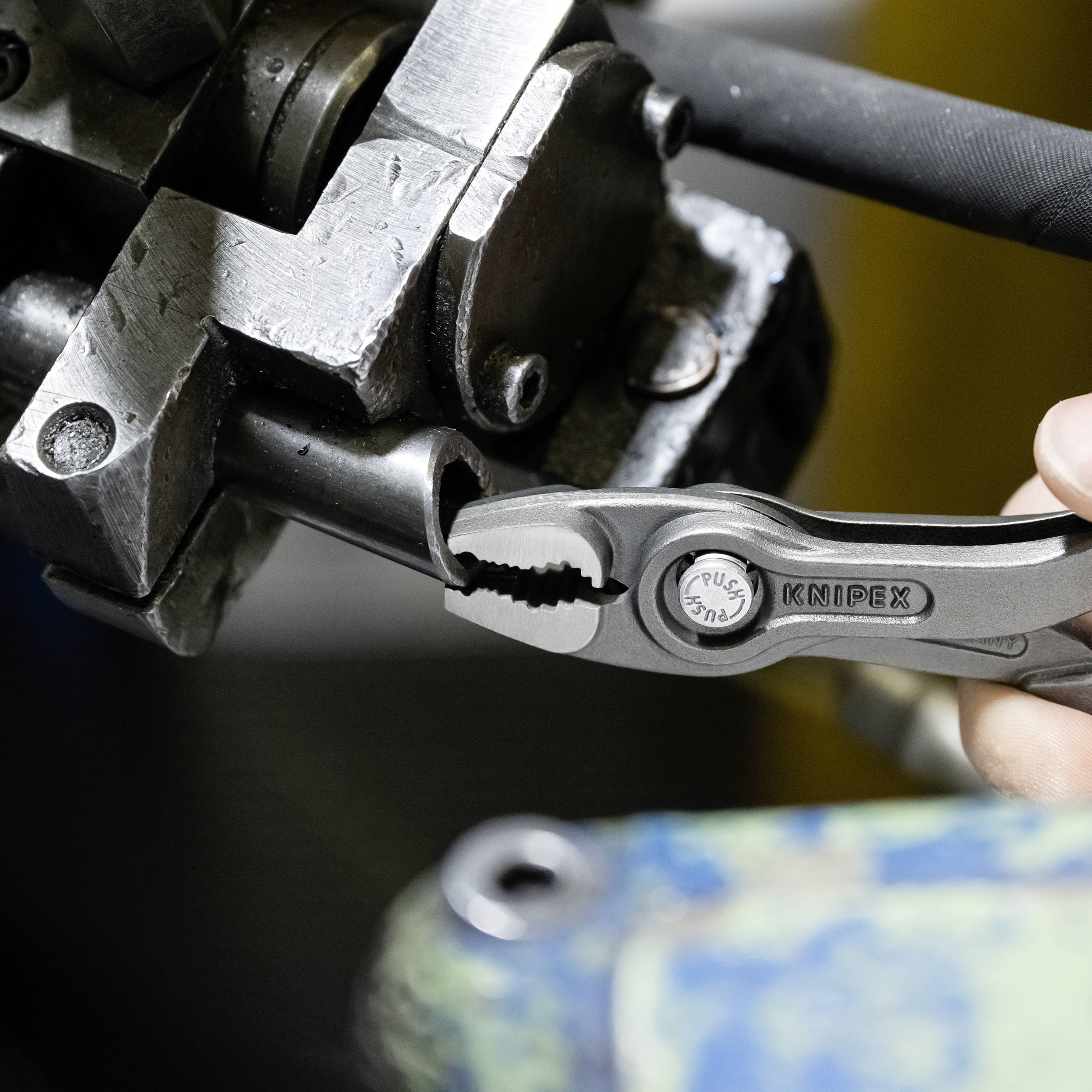 Egy kéz egy KNIPEX fogót tart és egy fémdarabot fog meg egy műhelykörnyezetben.