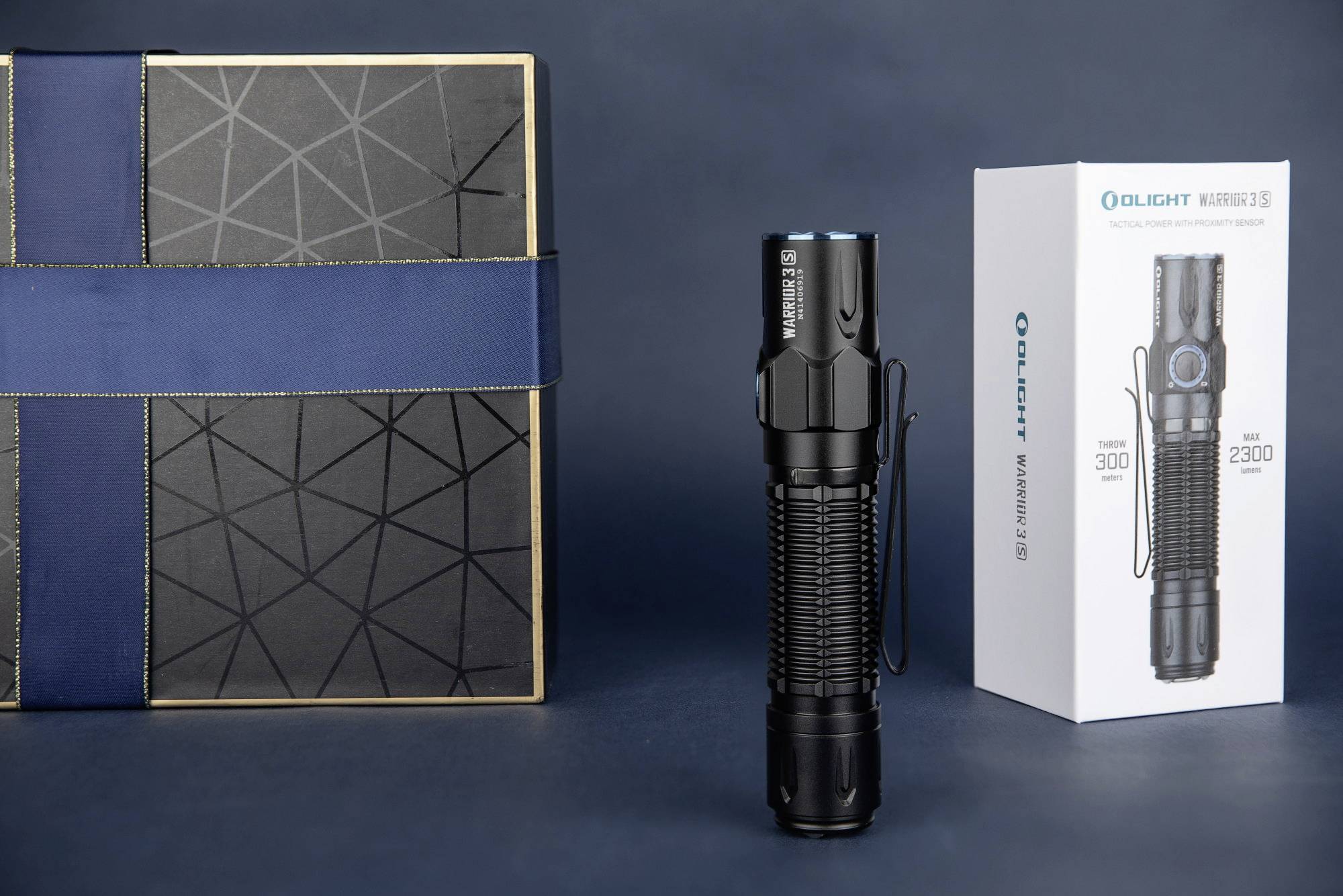 Egy fekete zseblámpa áll egyenesen egy dekoratív doboz és egy 'Olight Warrior 3S' feliratú csomag között.