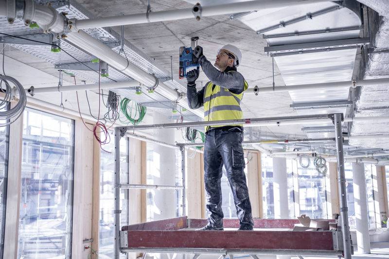 Egy munkás biztonsági mellényben és védősisakban áll egy állványzaton, és lyukakat fúr egy félig kész épület mennyezetébe.