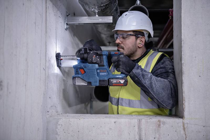 Egy munkás védősisakban és biztonsági mellényben fúr egy elektromos fúróval egy betonfalba.