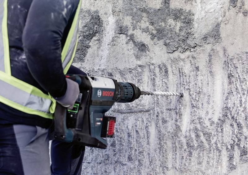 Egy munkásruhát viselő személy elektromos fúrógéppel lyukat fúr egy betonfalba. Biztonsági mellényt visel.