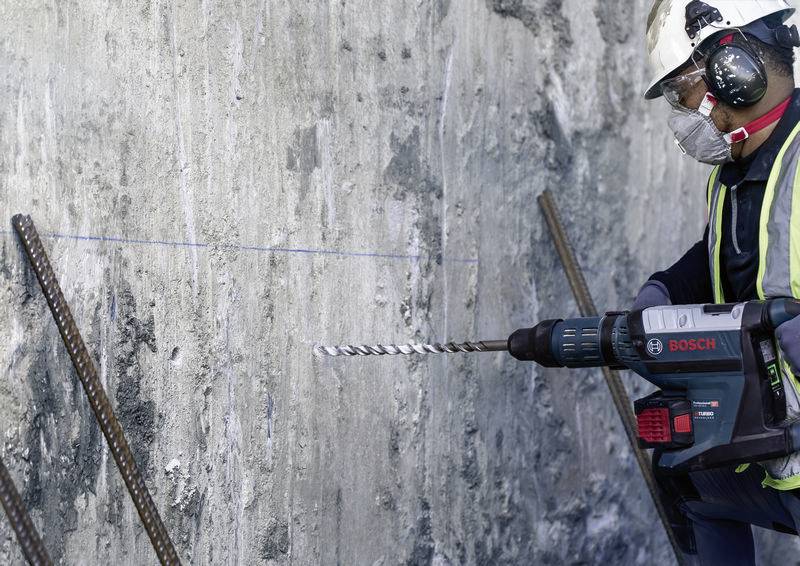 Egy munkás védőfelszerelésben és védősisakban elektromos fúrógéppel fúr egy betonfalat egy épülő munkaterületen.