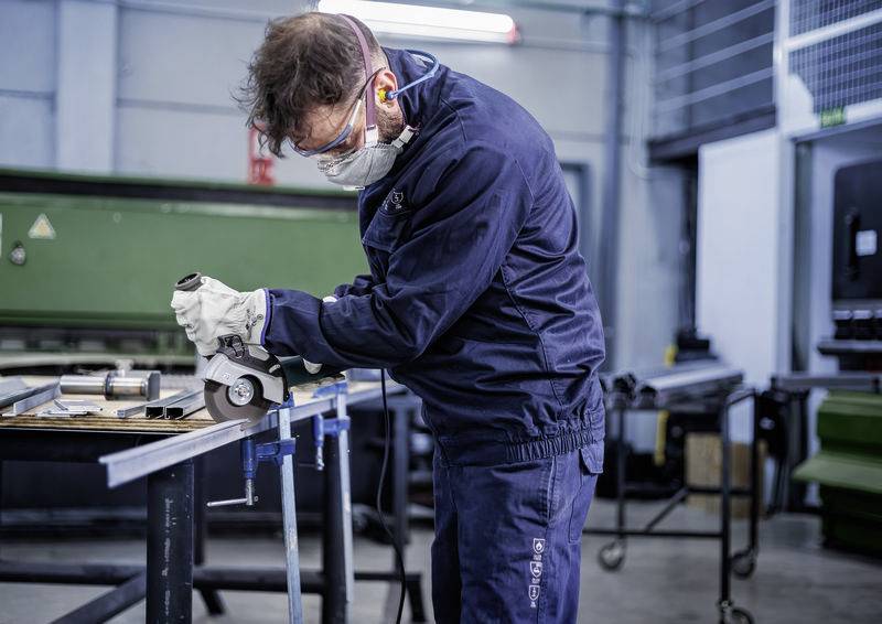 Egy férfi munkaruhában csiszol egy fémet egy műhelyben. Védőszemüveget, fülvédőt és biztonsági maszkot visel.
