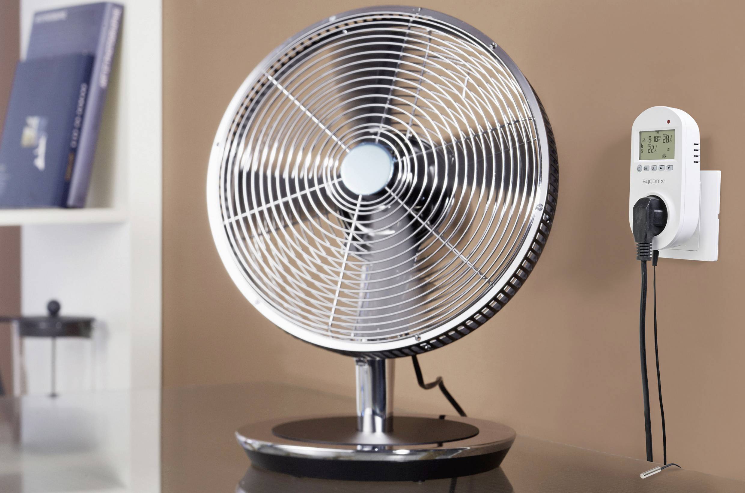 Egy asztali ventilátor fémráccsal áll az asztalon. Tőle jobbra egy falra szerelt konnektoros időkapcsoló látható.