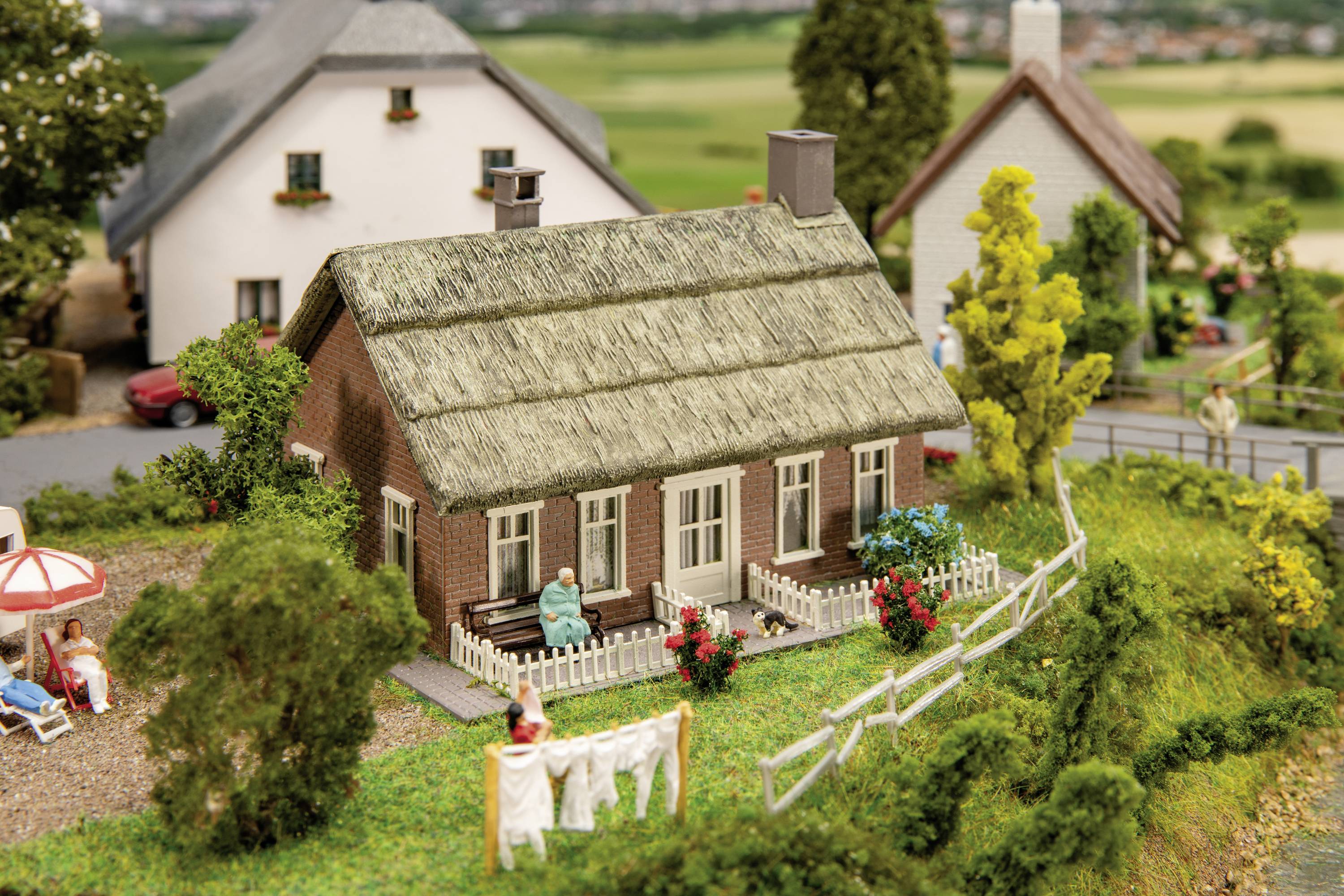 Egy falusi ház miniatűr modellje szalmatetővel, fák és kerítés között. Egy személy ül a teraszon.