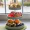 Egy háromszintes gyümölcstartó áll az asztalon, különböző gyümölcsökkel megrakva, mint alma, narancs és bogyók, egy ablak előtt.