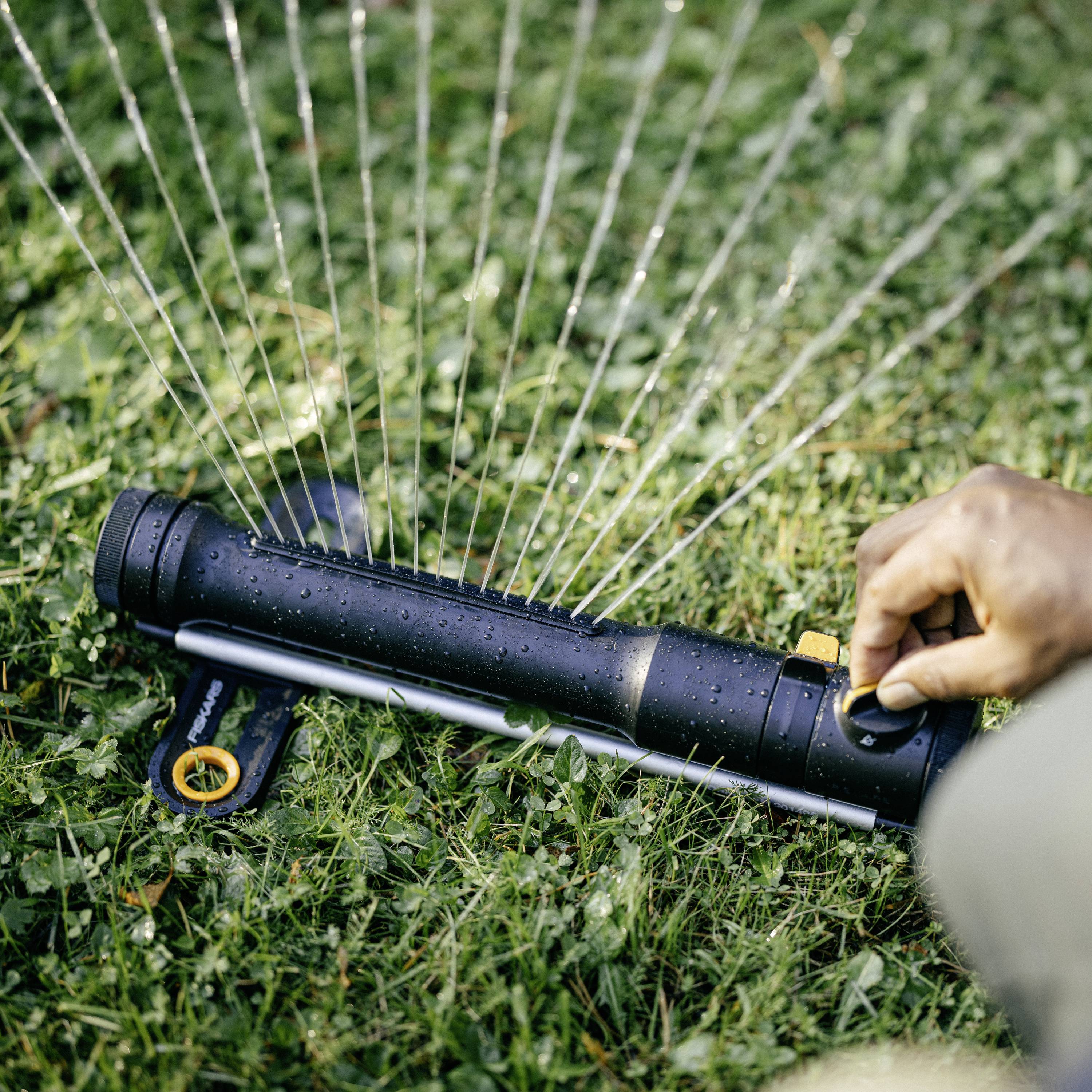 Egy zöld gyep fekete öntözőfejjel van bepermetezve vízzel, miközben egy kéz érinti az öntözőfejet.