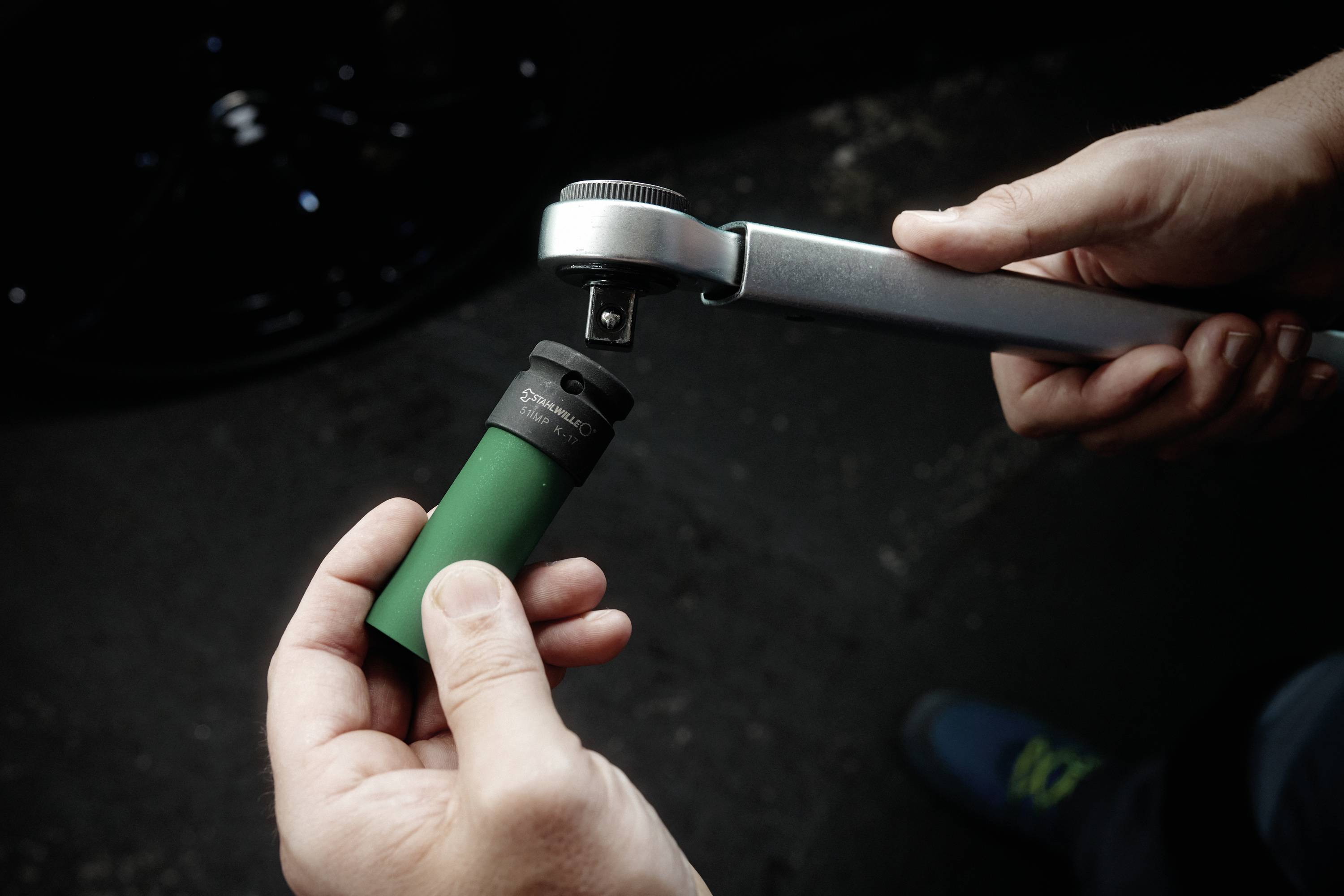 Egy személy kezei egy zöld dugófejű szerelvényt illeszt egy racsnis kulcsra, felkészítve azt a használatra, valószínűleg egy mechanikai vagy autóipari környezetben.