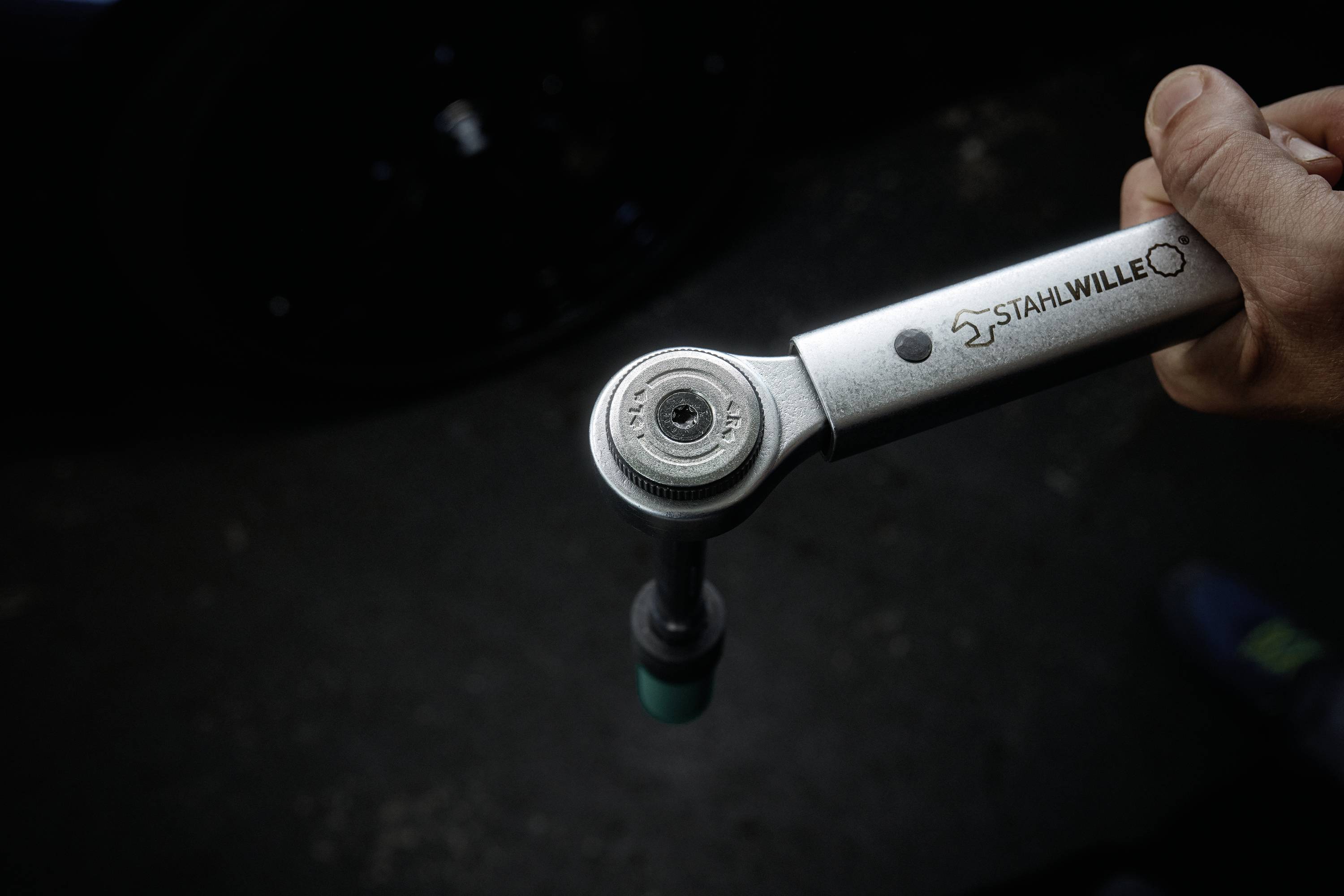 Egy kéz egy sötét felületen tart egy nyomatékkulcsot, amelyen a 'STAHLWILLE' felirat látható, miközben látszanak a szerszám vége és beállítási mechanizmusa.