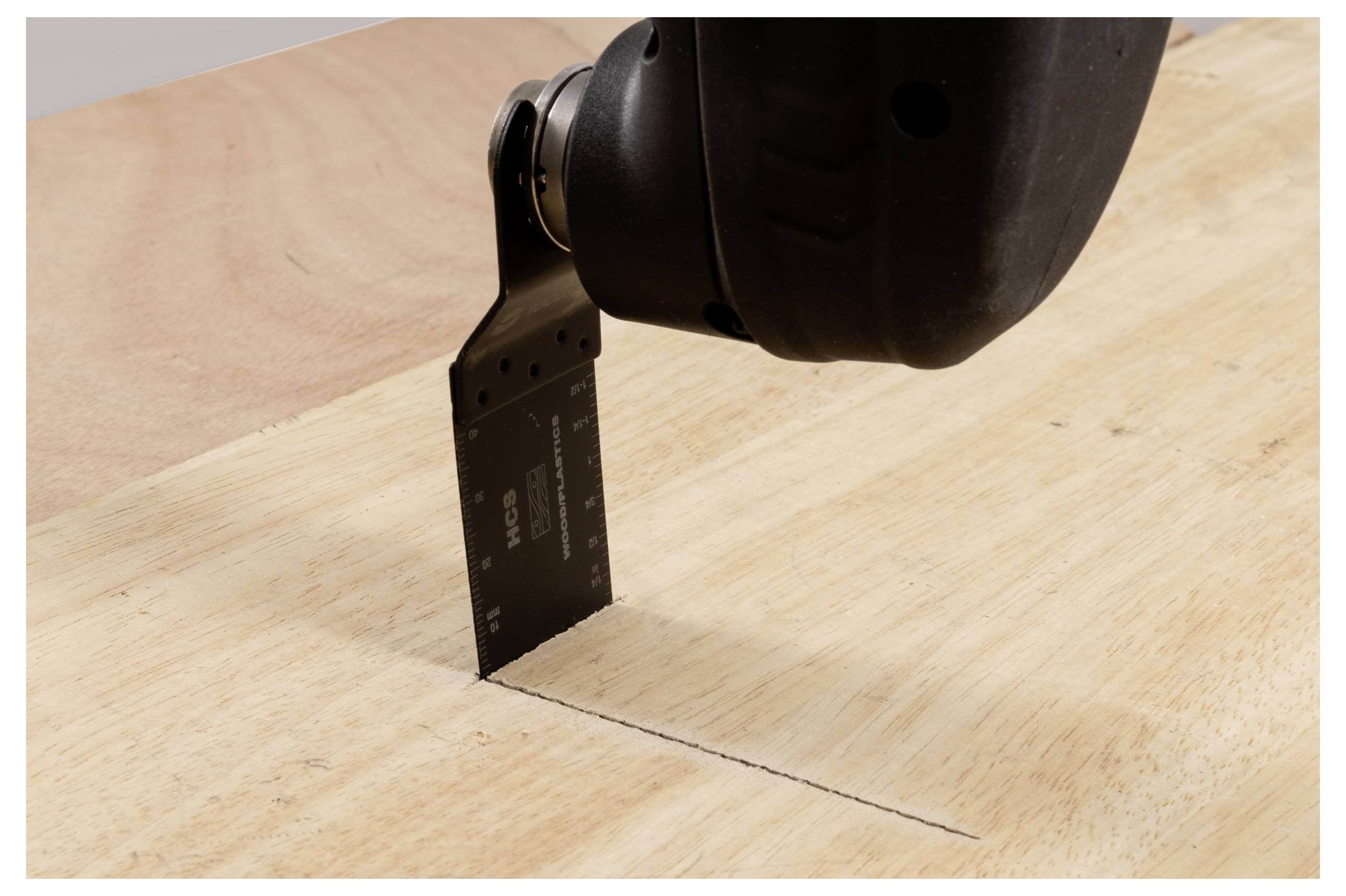 Egy elektromos kéziszerszám lapos pengével vág egy fadeszkába, egyenes vonalat húzva. Az eszköz lefelé döntött szögben van, precíz vágást mutatva.