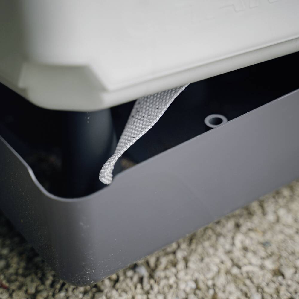 Egy szürke tartály öntözőkannával folyadékot ürít egy alatta elhelyezett felfogótálcába, kavicsokkal borított felületen.