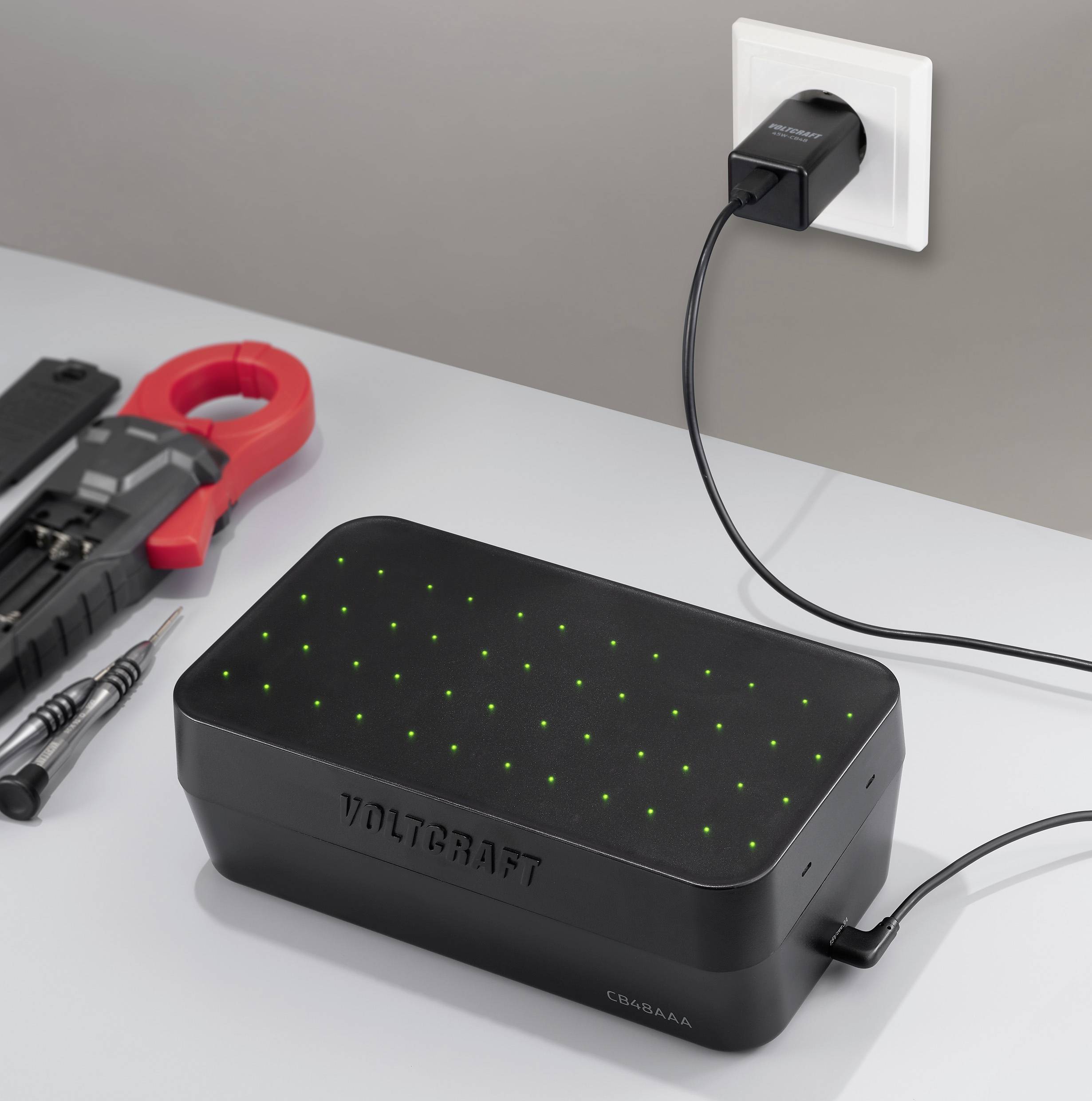 A 'Voltcraft' akkumulátortöltő egy asztalon szerszámok közelében, csatlakoztatva egy konnektorhoz. A lámpák jelzik a töltés állapotát.