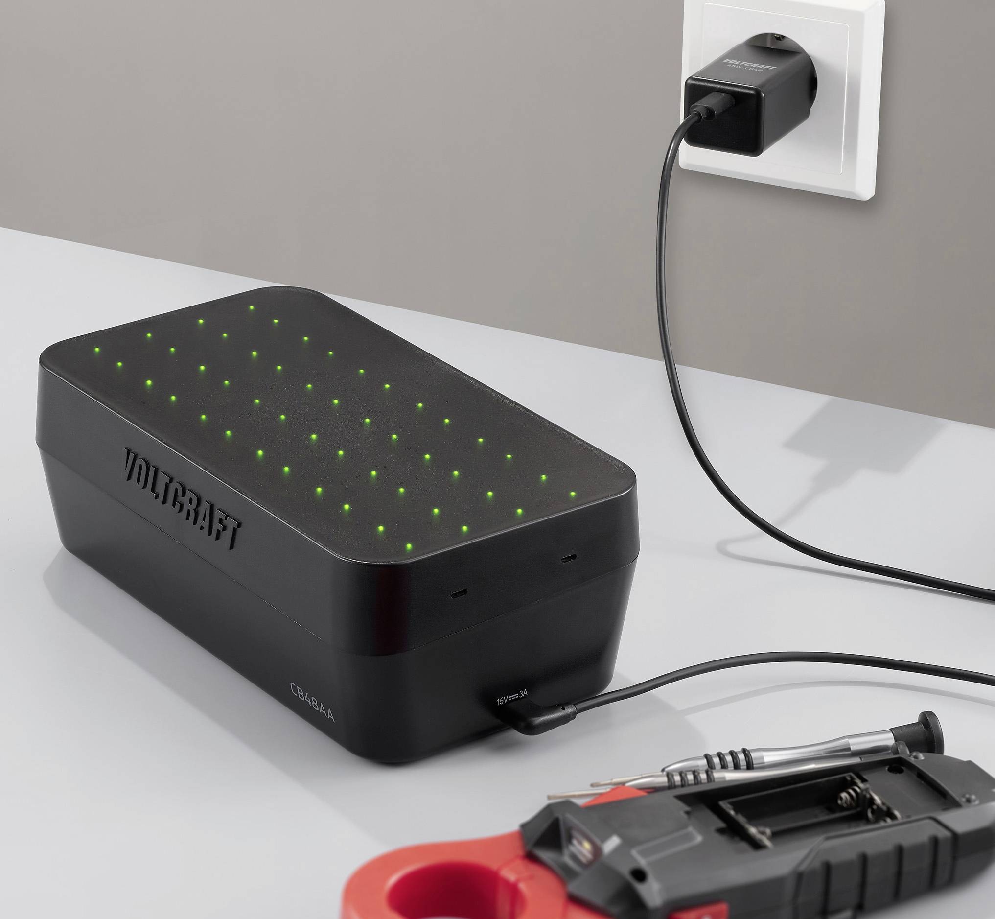 Egy fekete eszköz zöld LED-fényekkel egy fali konnektorba dugva, egy piros szerszámosláda és egy csavarhúzó mellett, szürke felületen.