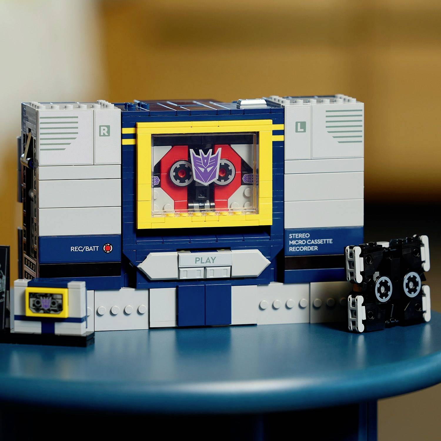 Egy játék kazettás lejátszó, amely egy robot fejére hasonlít, lila arccal és piros szemekkel, két kazettával az oldalán egy kék asztalon.