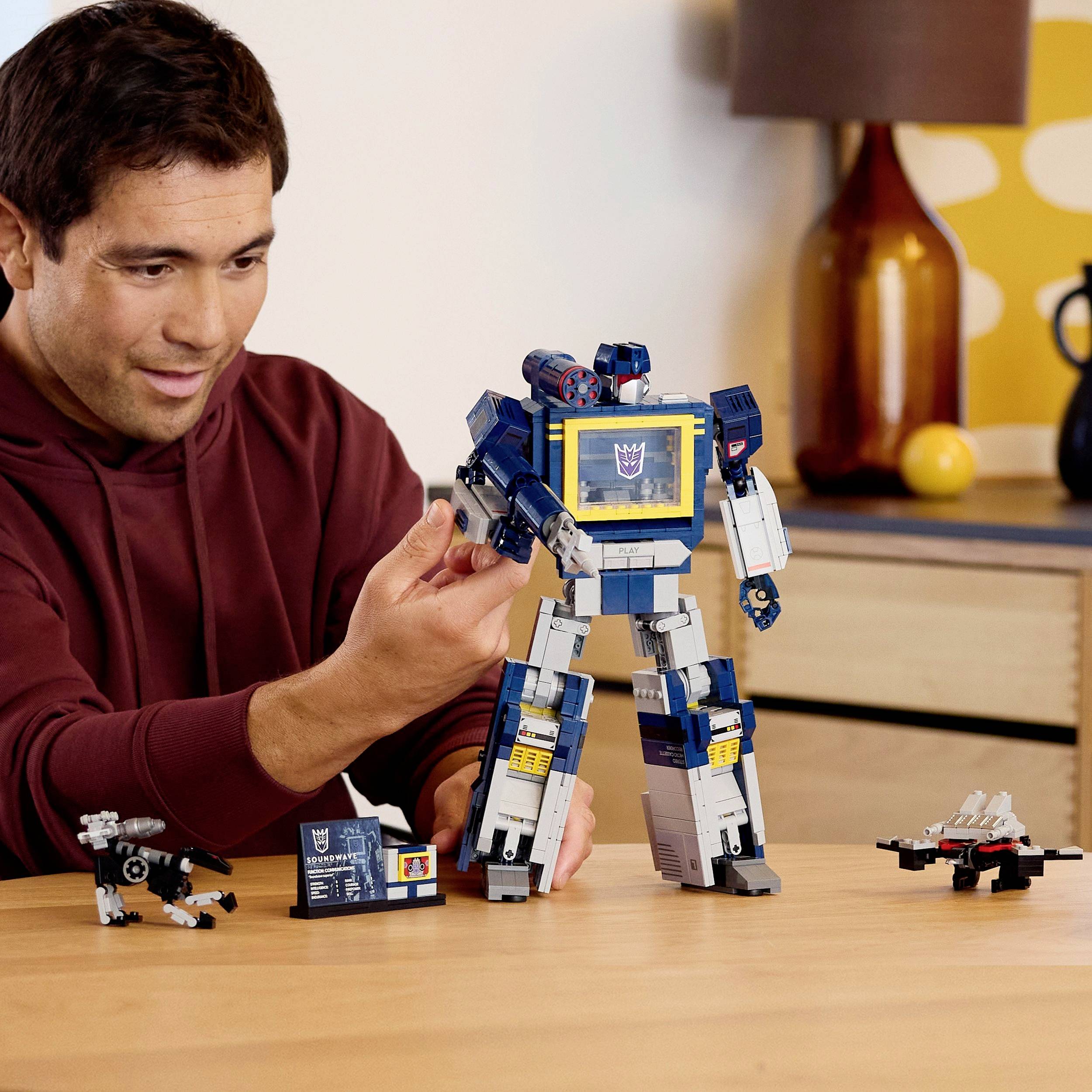 Egy személy egy nagy méretű robot játékkal kommunikál egy asztalon, kisebb robot figurák veszik körül, egy barátságos, meleg hangulatú szobában, amelyben egy fa komód is található.