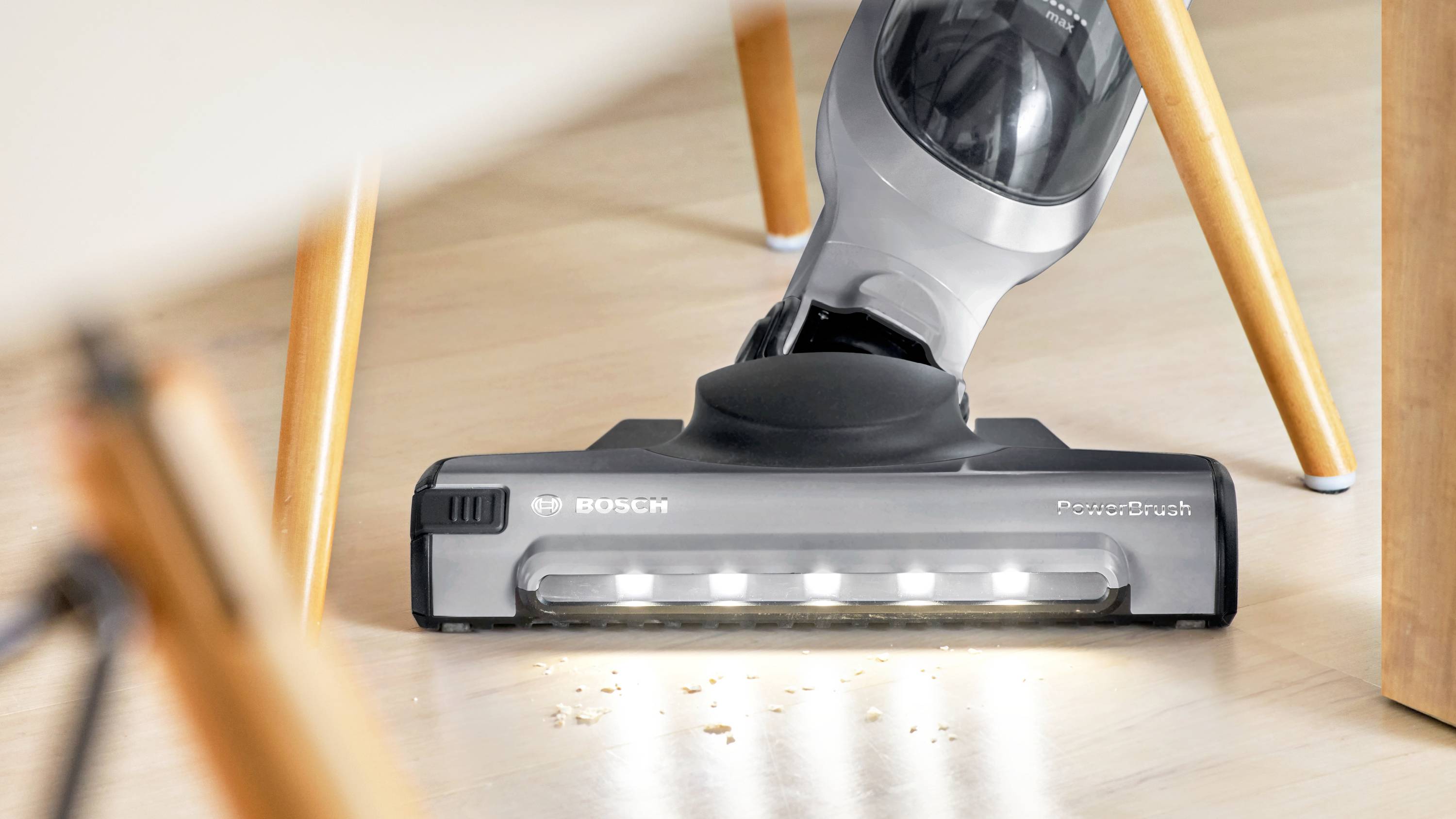 Egy LED-es lámpákkal ellátott porszívó hatékonyan takarítja fel a morzsákat egy sima, világos padlófelületről egy fa szék alatt.