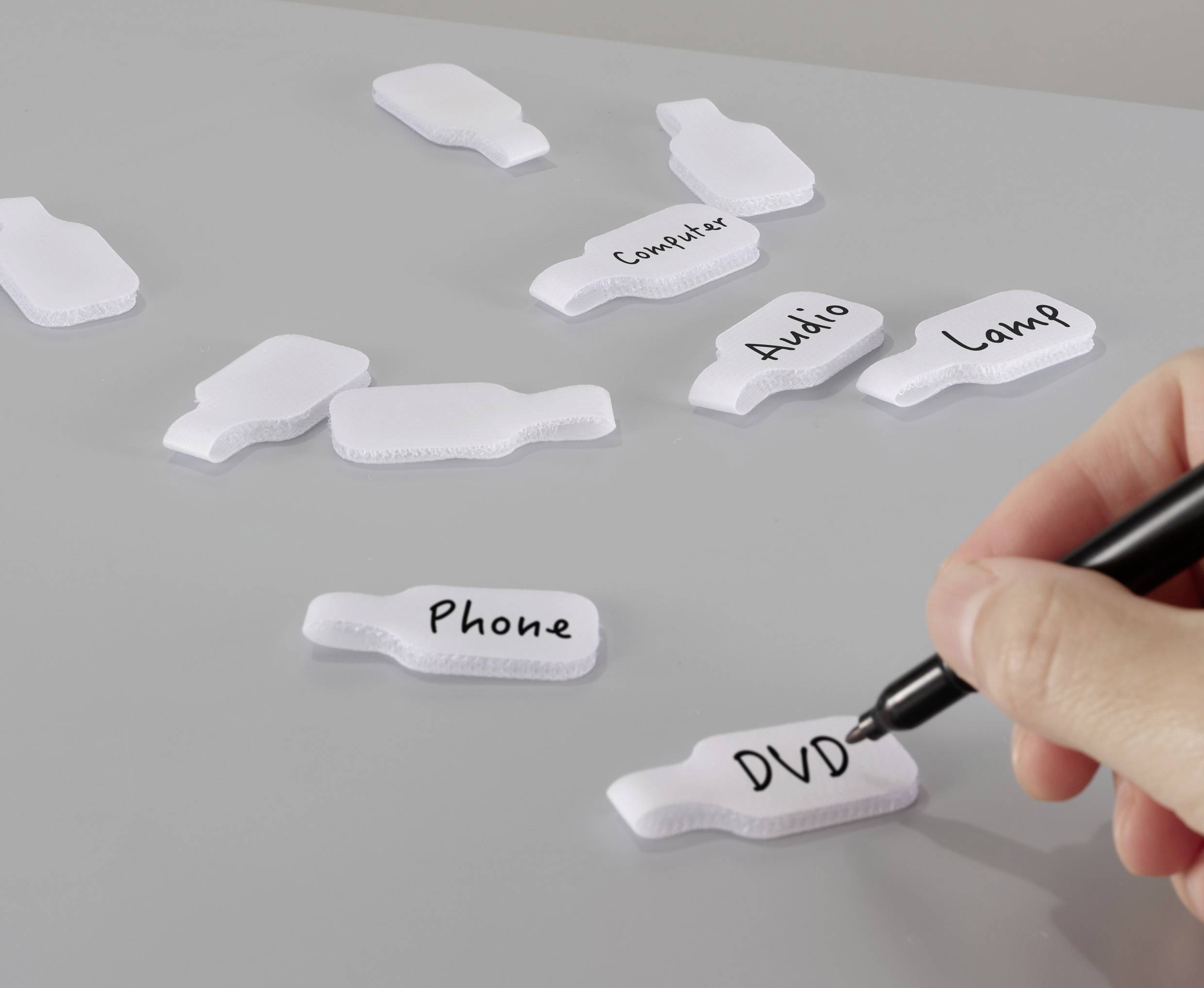 Egy kéz 'DVD' feliratot ír egy címkére, miközben más címkék olvashatók: 'Telefon', 'Számítógép', 'Audio', 'Lámpa', amelyek egy szürke háttéren szétszórva helyezkednek el.