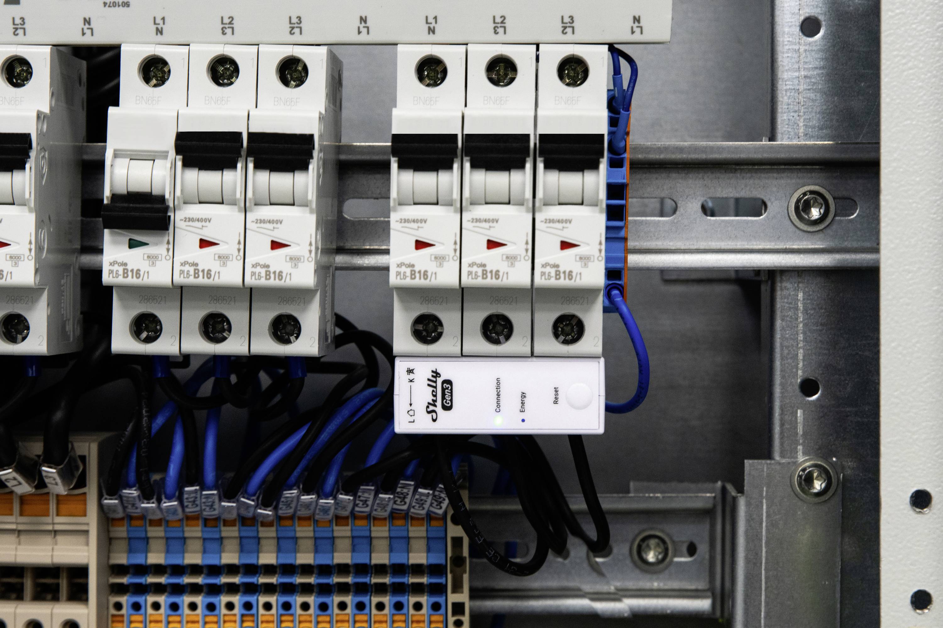 Egy elektromos elosztószekrény több áramköri megszakítóval és csatlakoztatott kék vezetékekkel. Egy fehér feszültségfigyelő eszköz jól láthatóan ki van emelve.