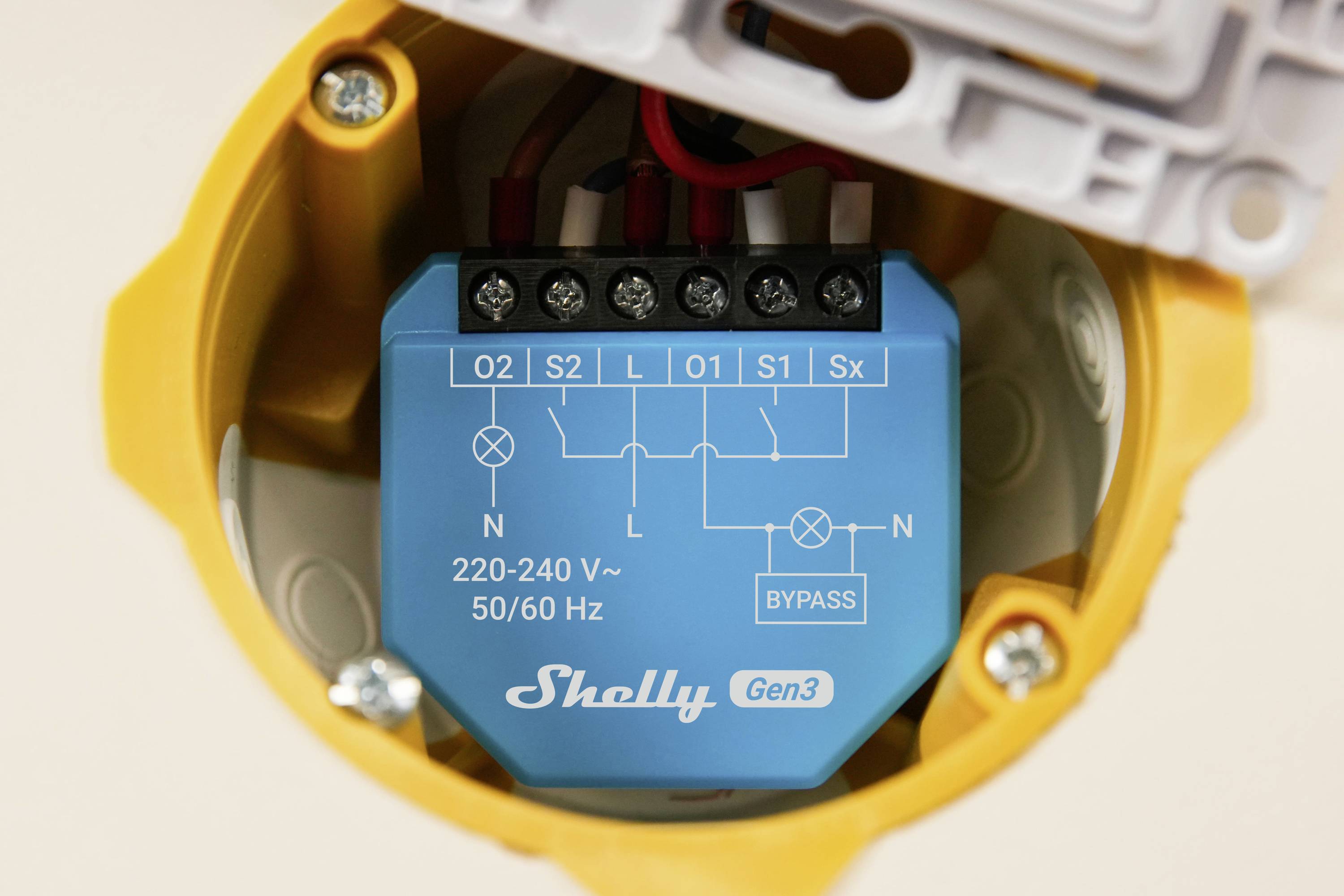 Egy kék Shelly Gen3 intelligens kapcsoló egy elektromos dobozban, amelyen láthatók a huzalozási csatlakozások az O2, S2, L, O1, S1 és nulla vezetékekhez. Megjelölve: '220-240 V~ 50/60 Hz'.
